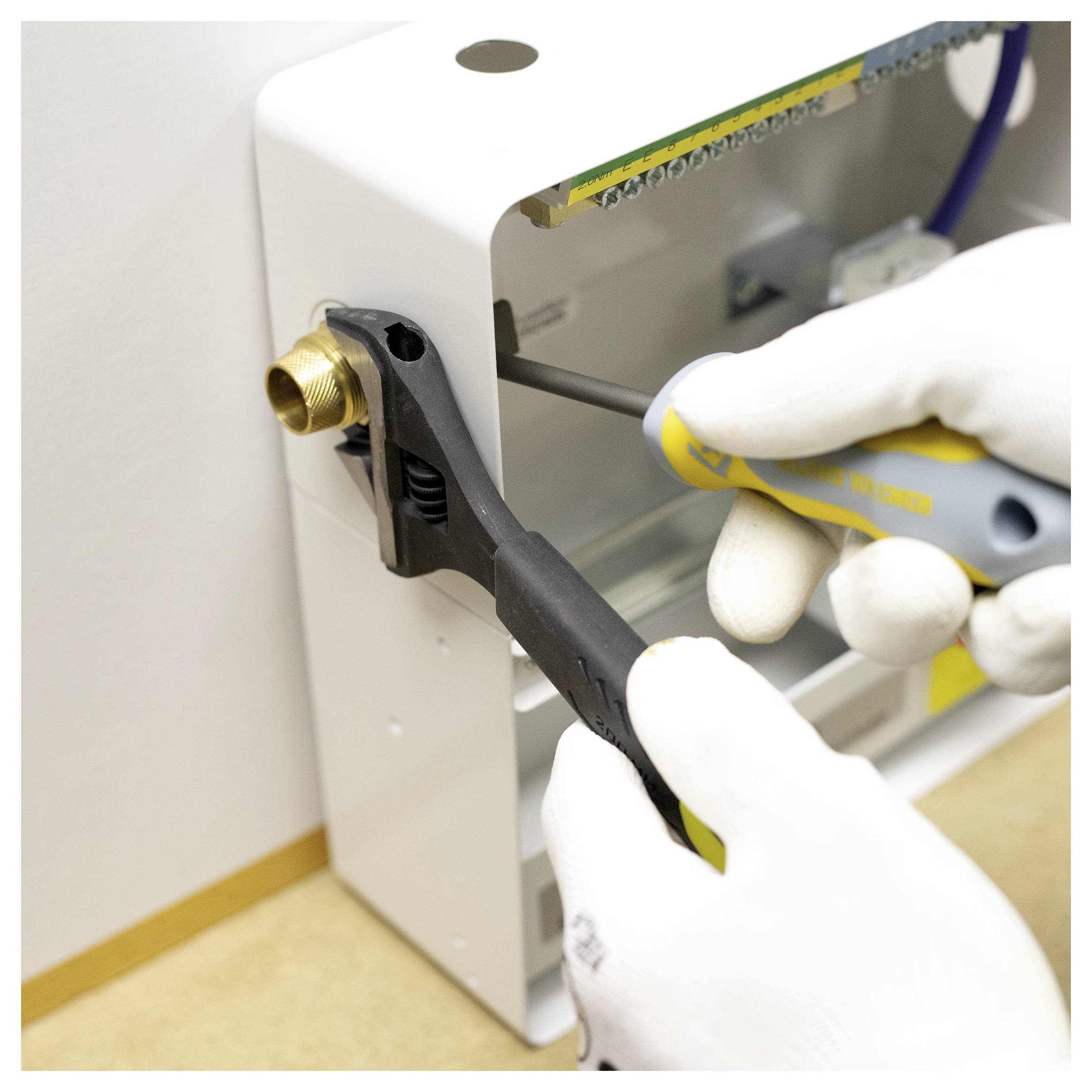 Eine Person, die Handschuhe trägt, benutzt einen Schraubenschlüssel, um eine Messingmutter an einem weißen Industriegerät einzustellen und demonstriert dabei ein Wartungsverfahren.