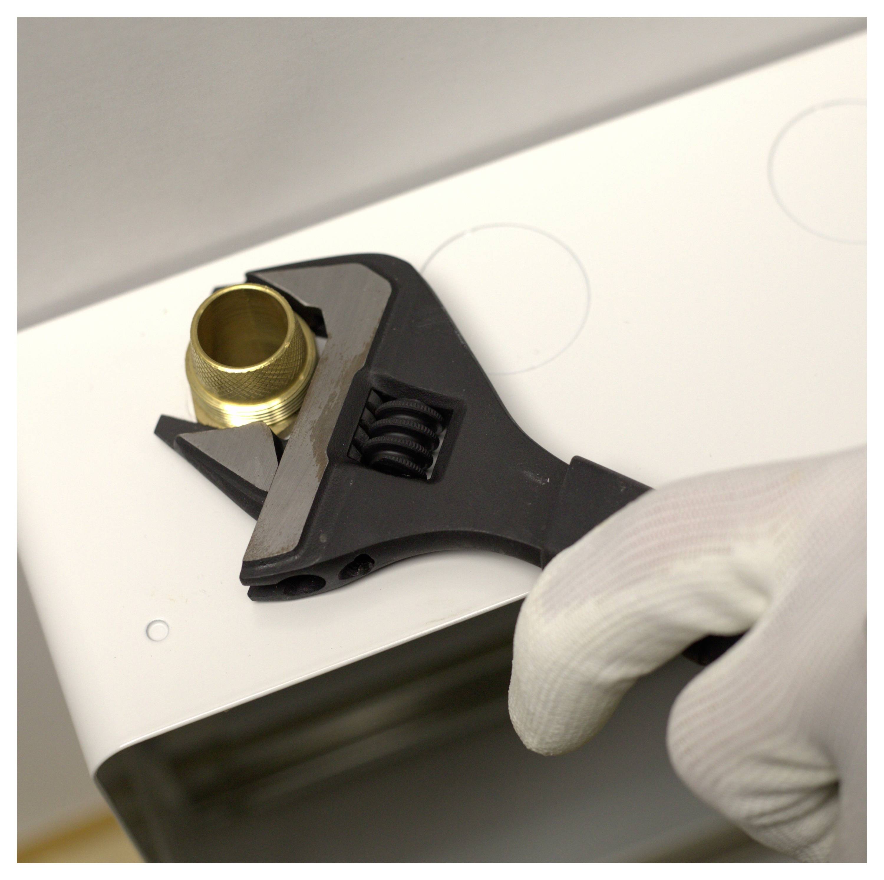 Eine behandschuhte Hand benutzt einen verstellbaren Schraubenschlüssel, um eine Messingarmatur auf einer weißen Oberfläche festzuziehen, möglicherweise im Zusammenhang mit Klempnerarbeiten oder Montagearbeiten.