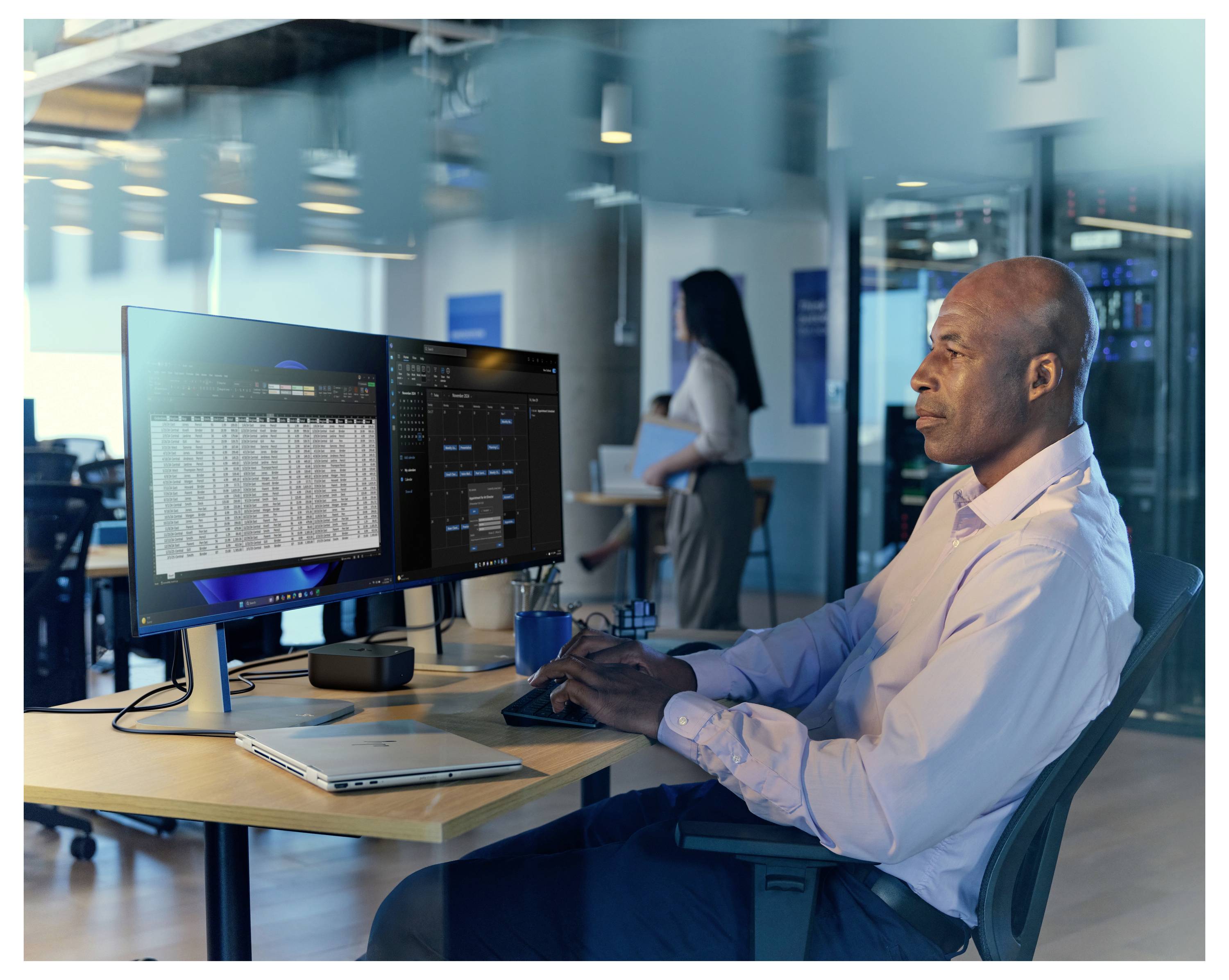 Eine Person in einem Videoanruf mit mehreren Teilnehmern, die auf einem Computermonitor angezeigt werden, während Tabellenkalkulationsdaten auf einem anderen Bildschirm sichtbar sind.