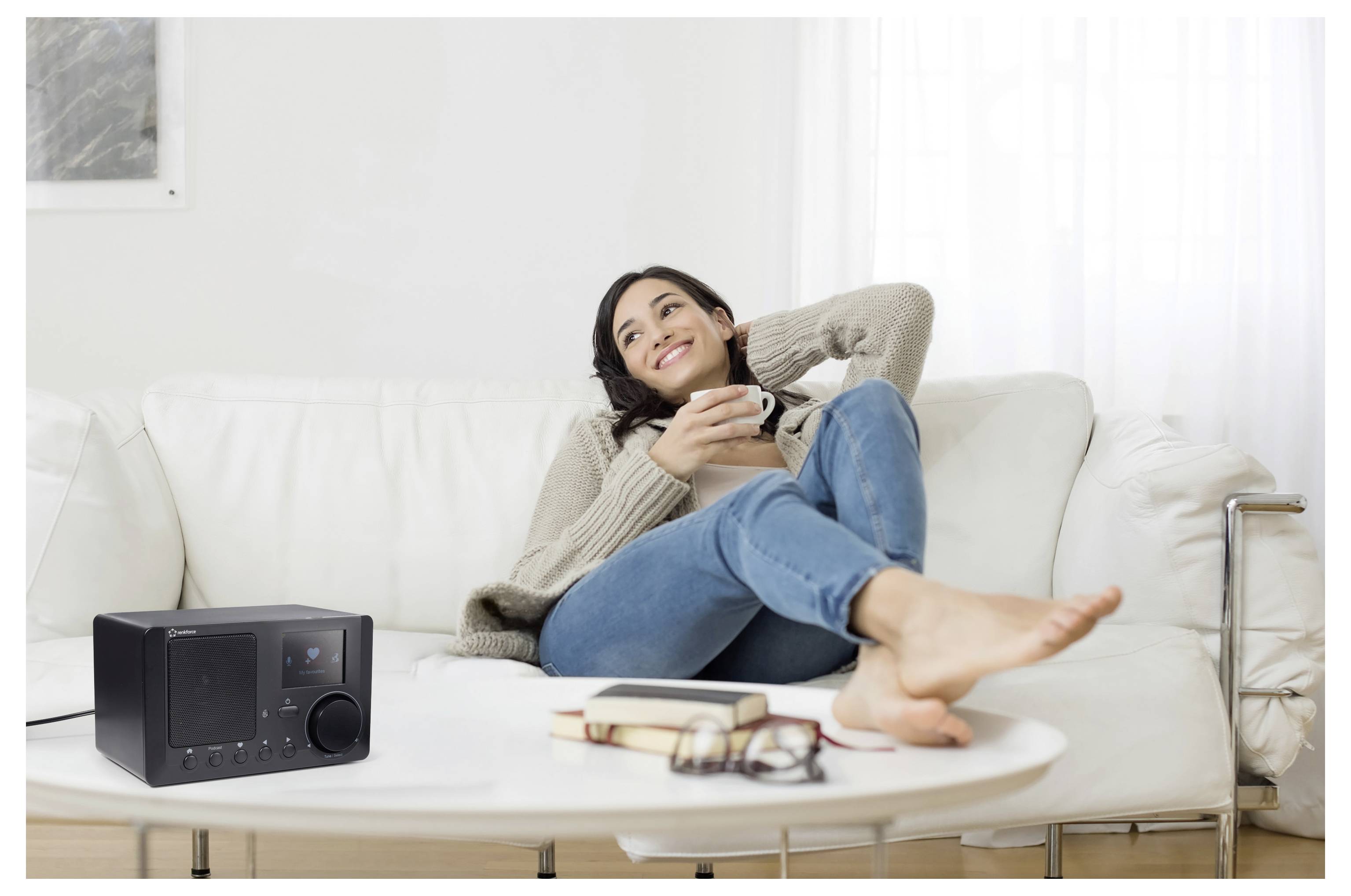 Eine Frau, die sich auf einem weißen Sofa entspannt, lächelt während sie eine Tasse hält; ein Radio sitzt auf dem Tisch neben Büchern und einer Brille.