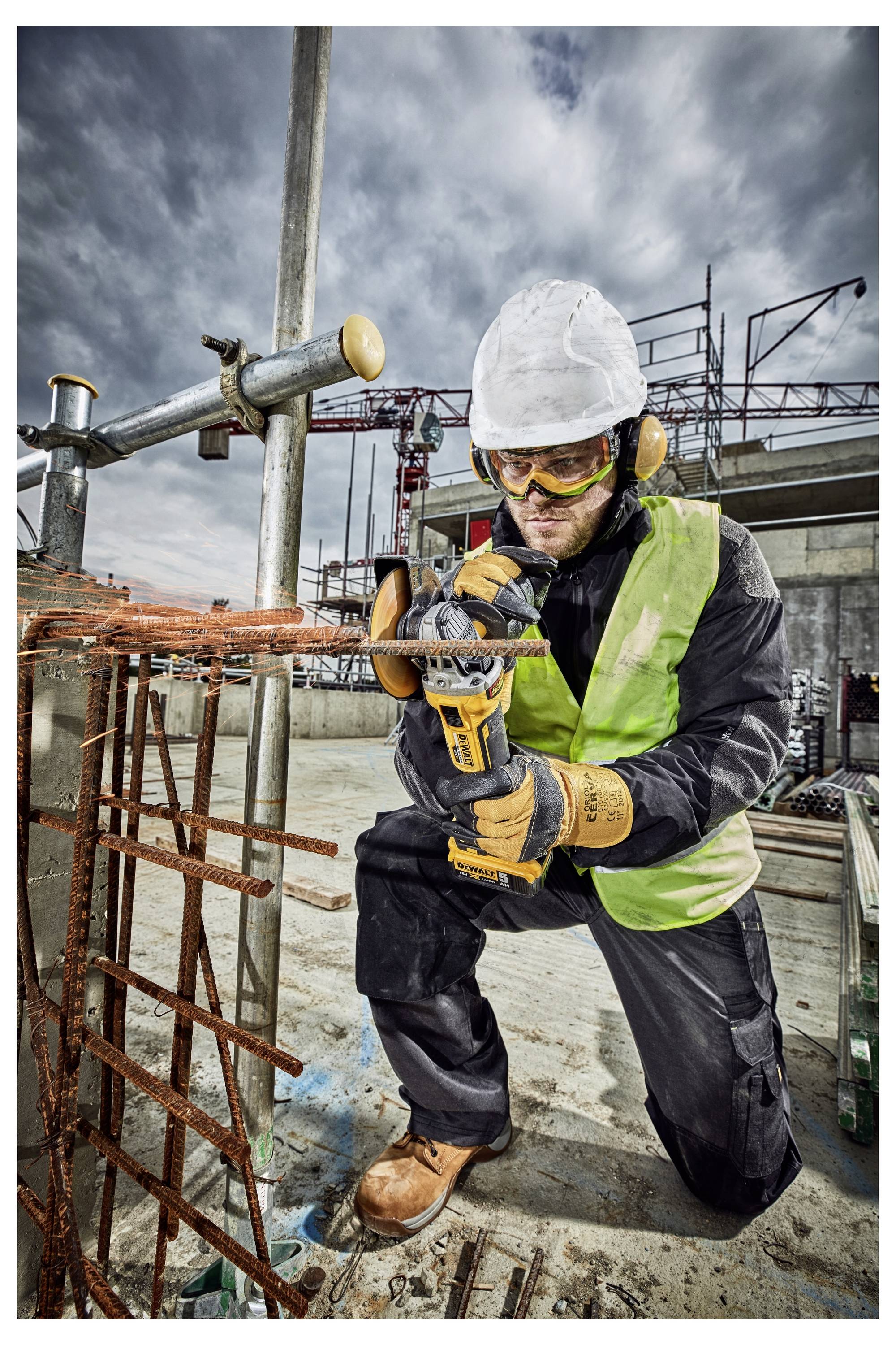 Ein Bauarbeiter in Schutzausrüstung verwendet ein Elektrowerkzeug zum Schneiden von Metallbewehrung auf einer Baustelle, mit Gerüsten und bewölktem Himmel im Hintergrund.