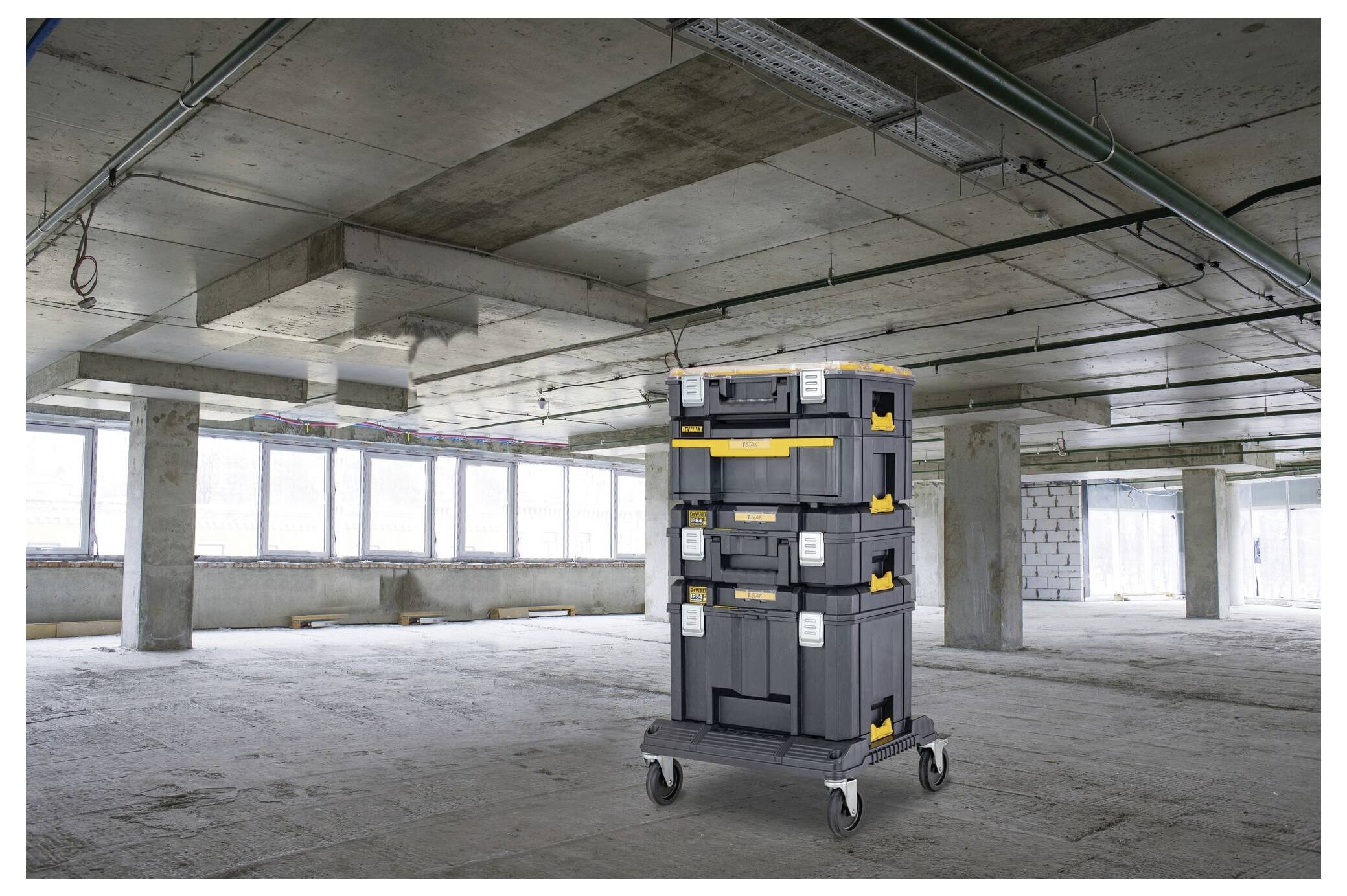 Drei übereinander gestapelte Lagerkästen auf Rollen in einem unvollendeten Betonraum mit großen Fenstern, Rohren und unvollendeten Wänden.