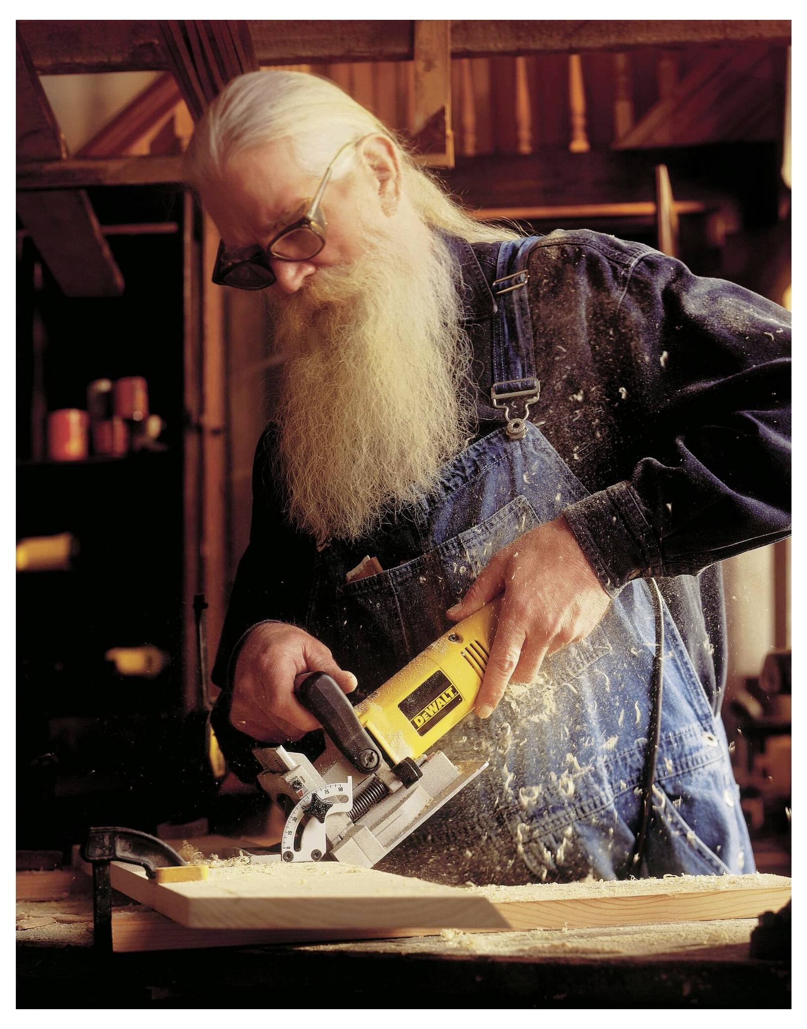Eine Person mit langen Haaren und einem Bart, die eine Sonnenbrille und einen Jeansoverall trägt, benutzt eine gelbe Elektrosäge an einem Holzstück in einer Werkstatt.