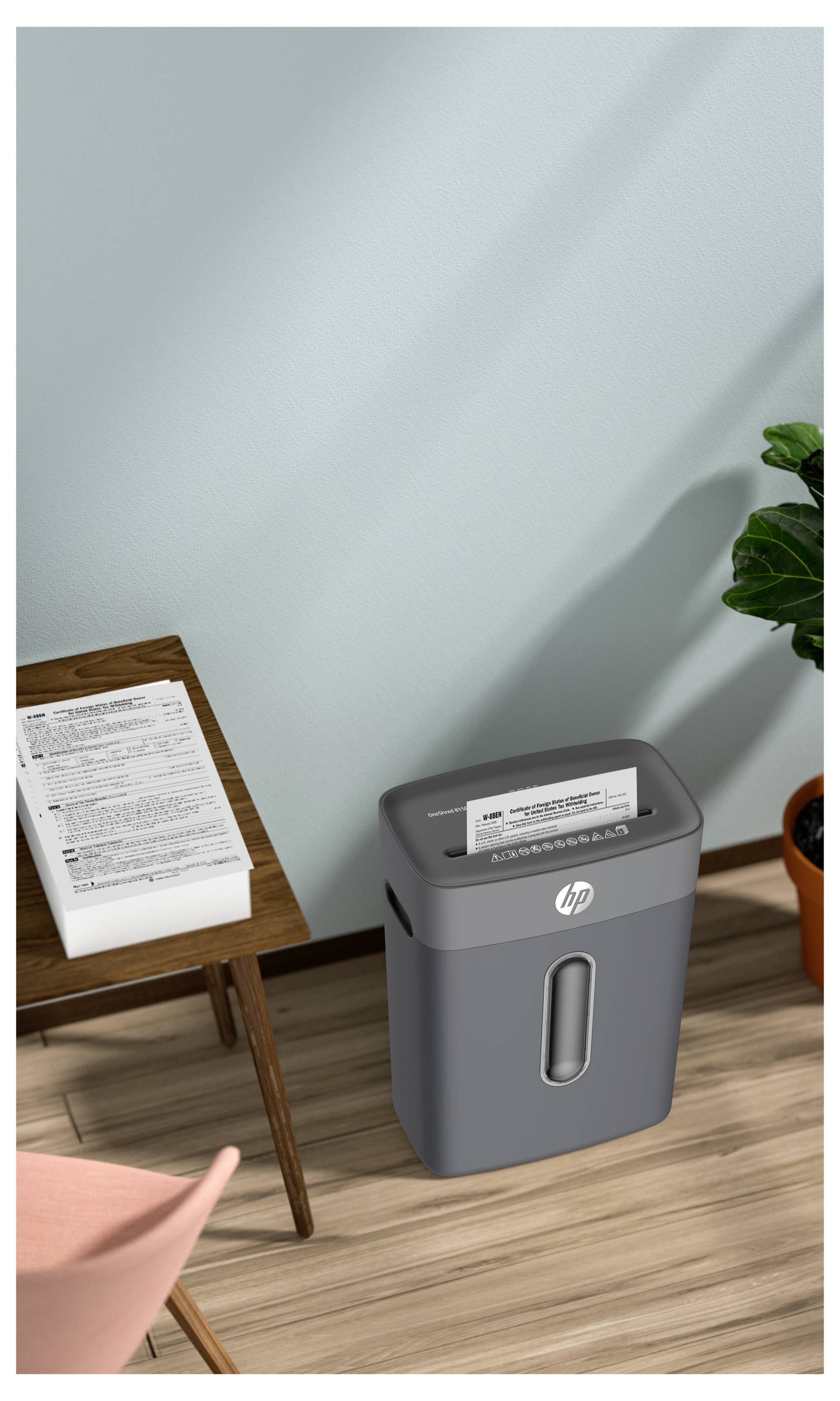 Ein Dokumentenschredder neben einem Stapel Papiere auf einem Holztisch. Eine Pflanze in einem Topf steht in der Nähe, alles vor einer hellblauen Wand.