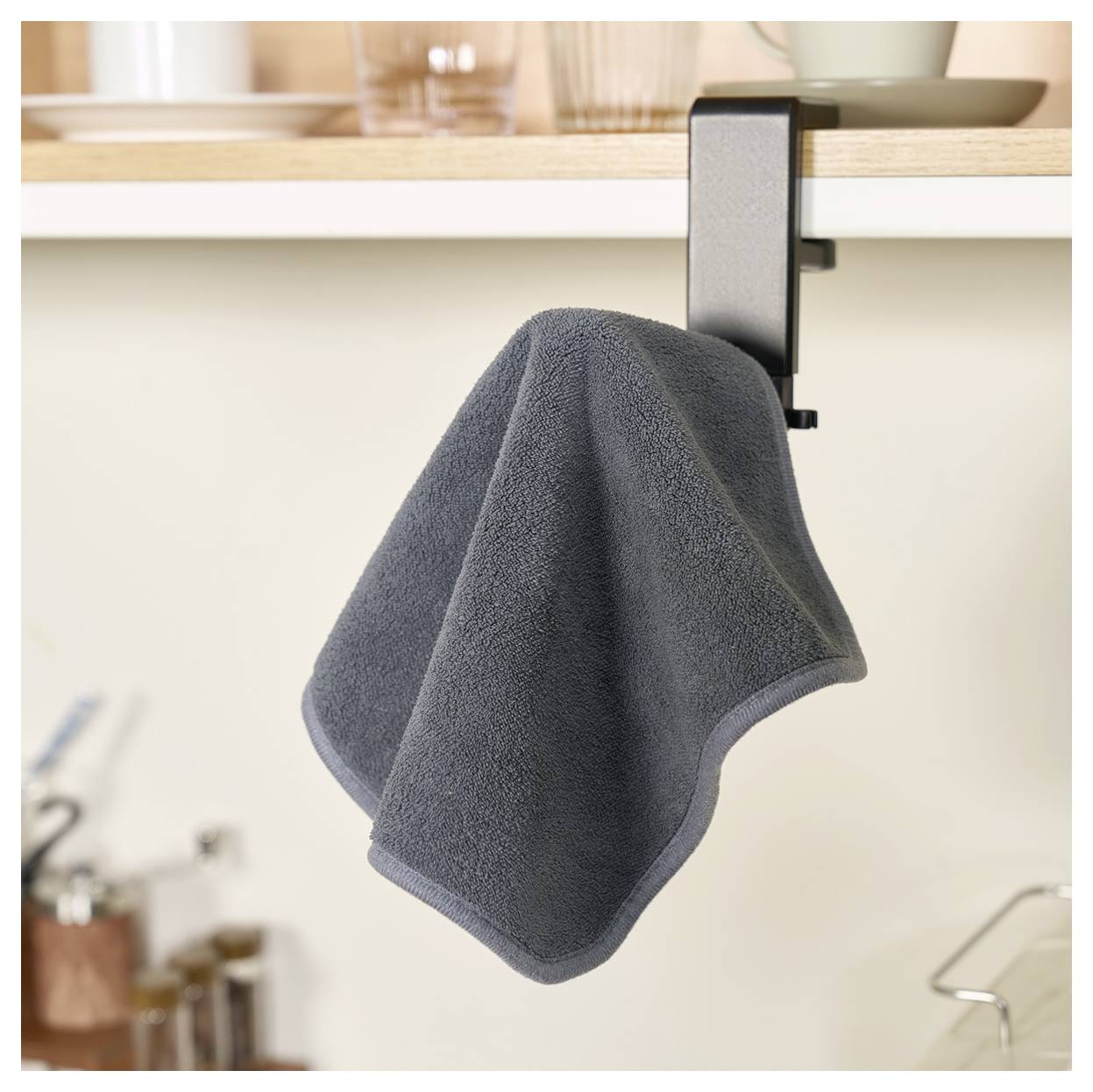 Ein schwarzes Geschirrtuch, das an einem Haken unter einem Küchenregal hängt, mit unscharfen Küchenutensilien im Hintergrund.