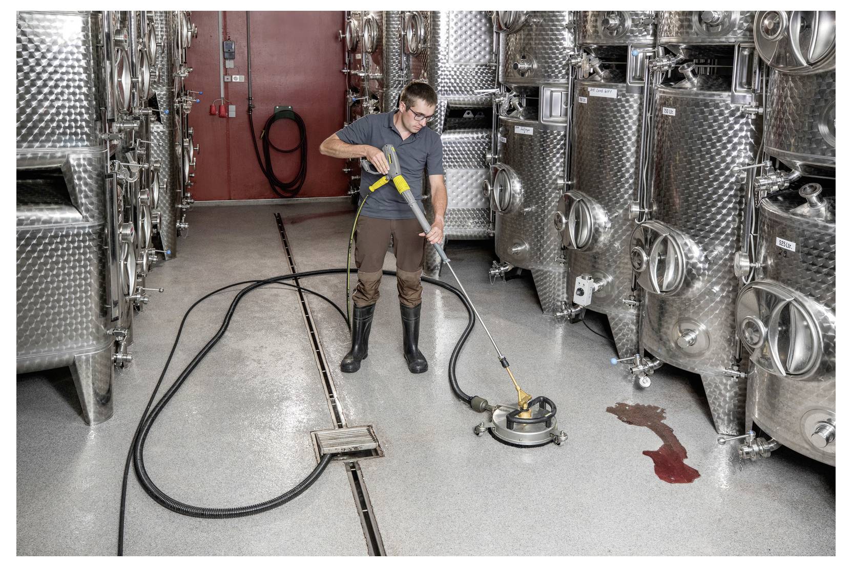 Eine Person in Stiefeln bedient eine Bodentansteinigung in einer industriellen Umgebung mit Metalltanks. In der Nähe befindet sich eine rote Flüssigkeitsverschüttung.