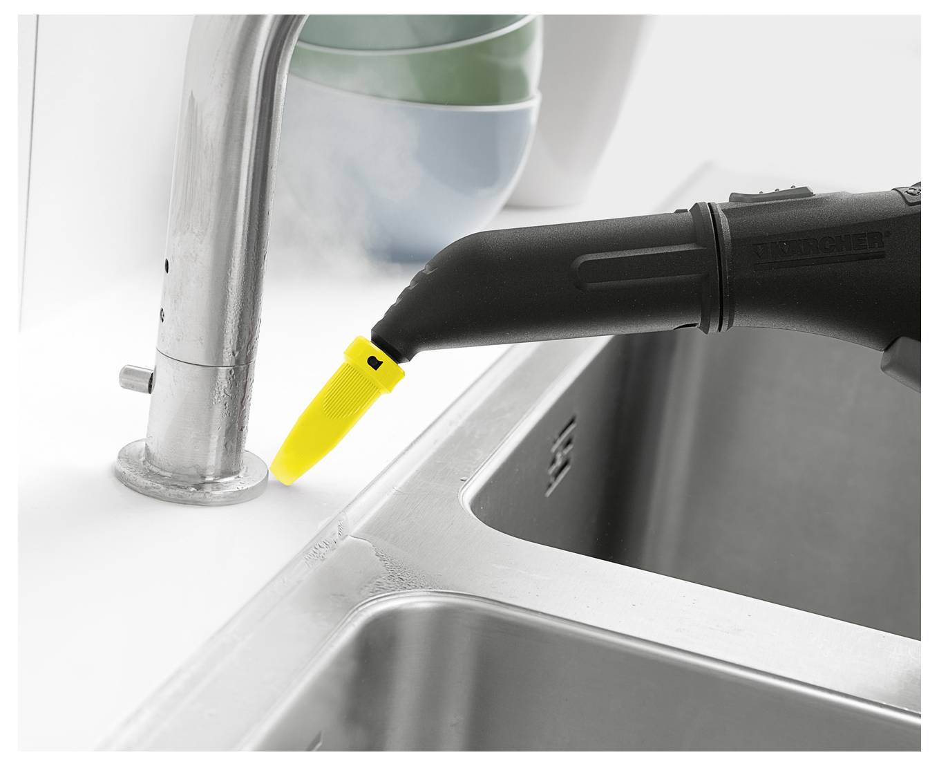 Eine Dampfreinigungsdüse, die Dampf abgibt, während sie den Fuß eines Küchenspülbecken-Wasserhahns reinigt, mit einem Spülbecken und Geschirr im Hintergrund.