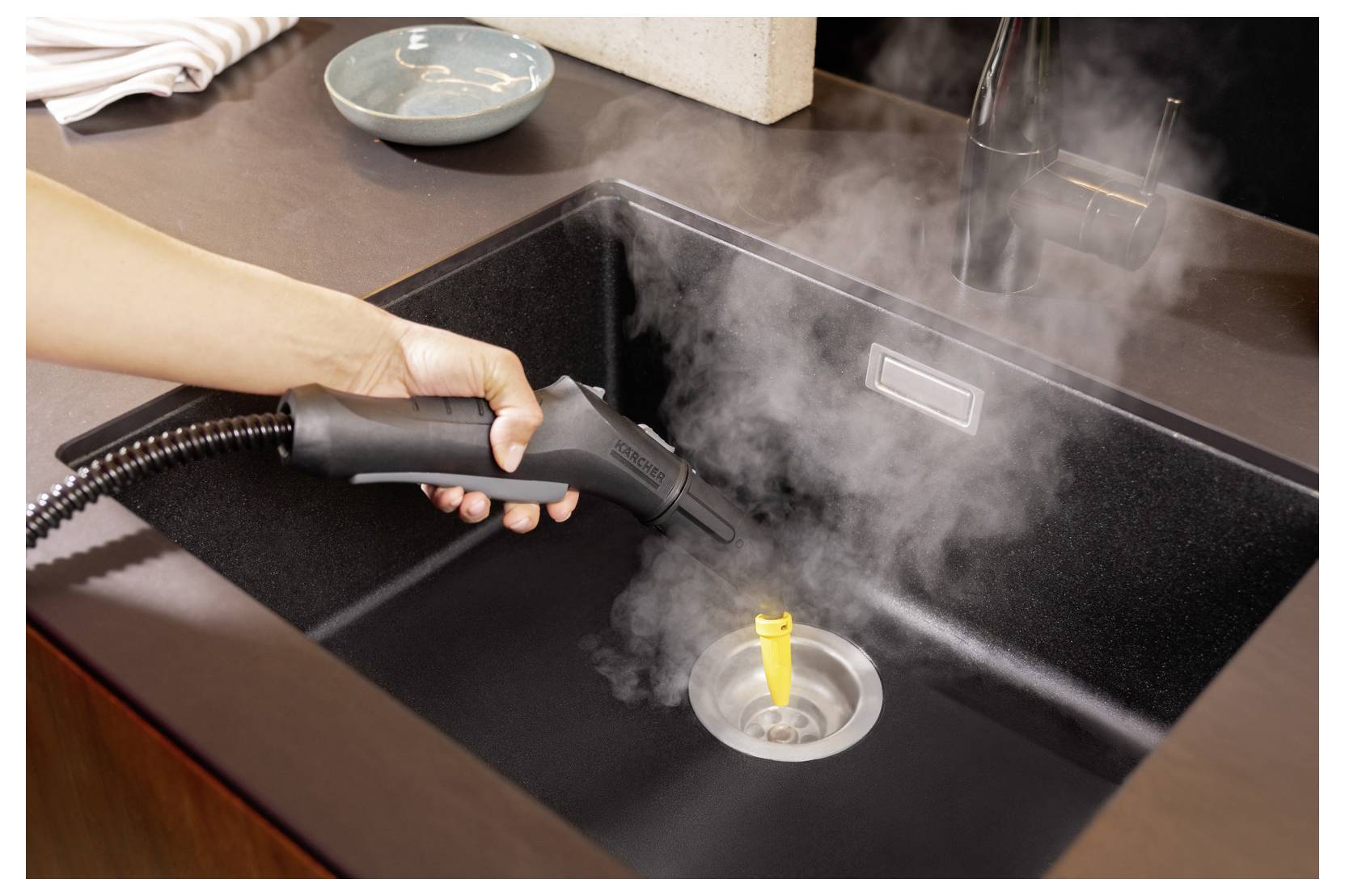 Eine Hand benutzt einen Dampfreiniger, um ein Küchenspülbecken zu desinfizieren, wobei Dampf deutlich aufsteigt.