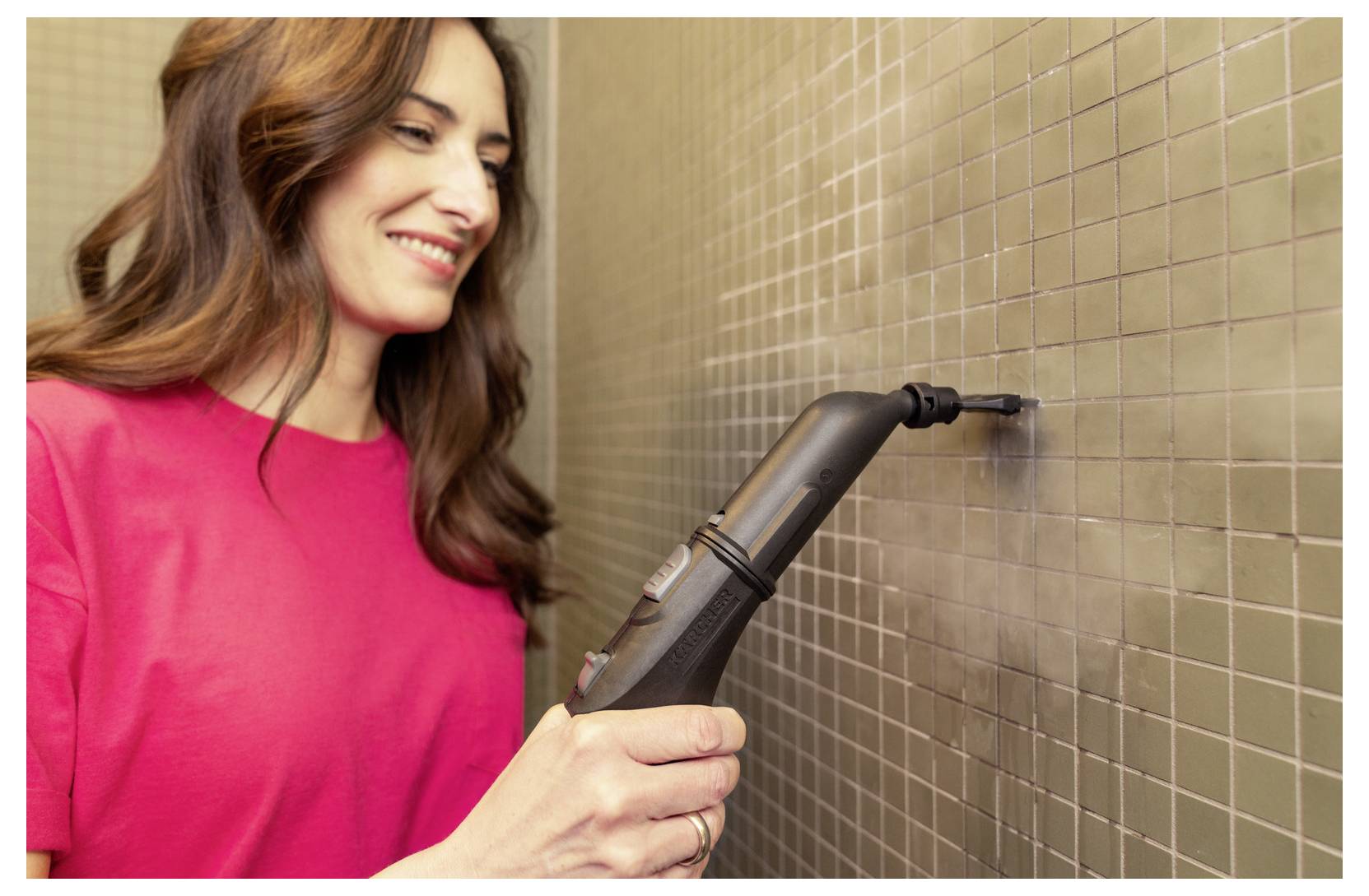 Eine Frau im roten Hemd benutzt einen Dampfreiniger an einer gekachelten Wand und lächelt dabei, während sie arbeitet.