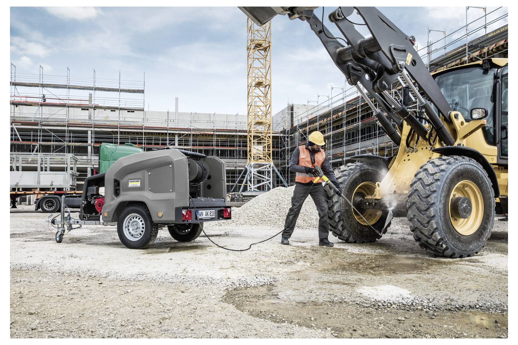 Ein Bauarbeiter nutzt einen Hochdruckreiniger, der an einen tragbaren Luftkompressor angeschlossen ist, um einen großen Fahrzeugreifen auf einer belebten Baustelle zu reinigen.