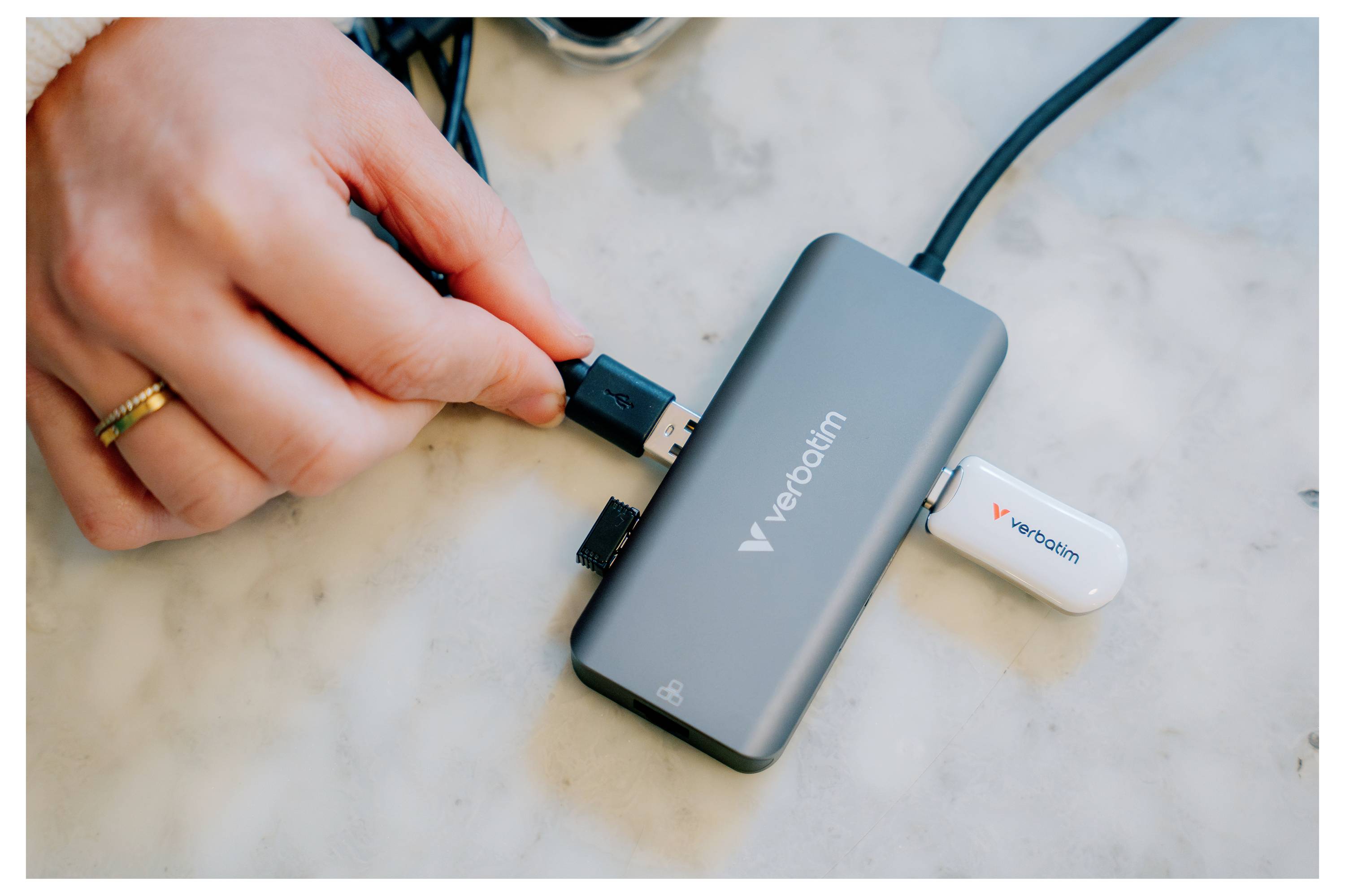 Hand steckt ein USB-Kabel in einen grauen Verbatim-Hub mit einem angeschlossenen weißen USB-Stick auf einer Marmoroberfläche.