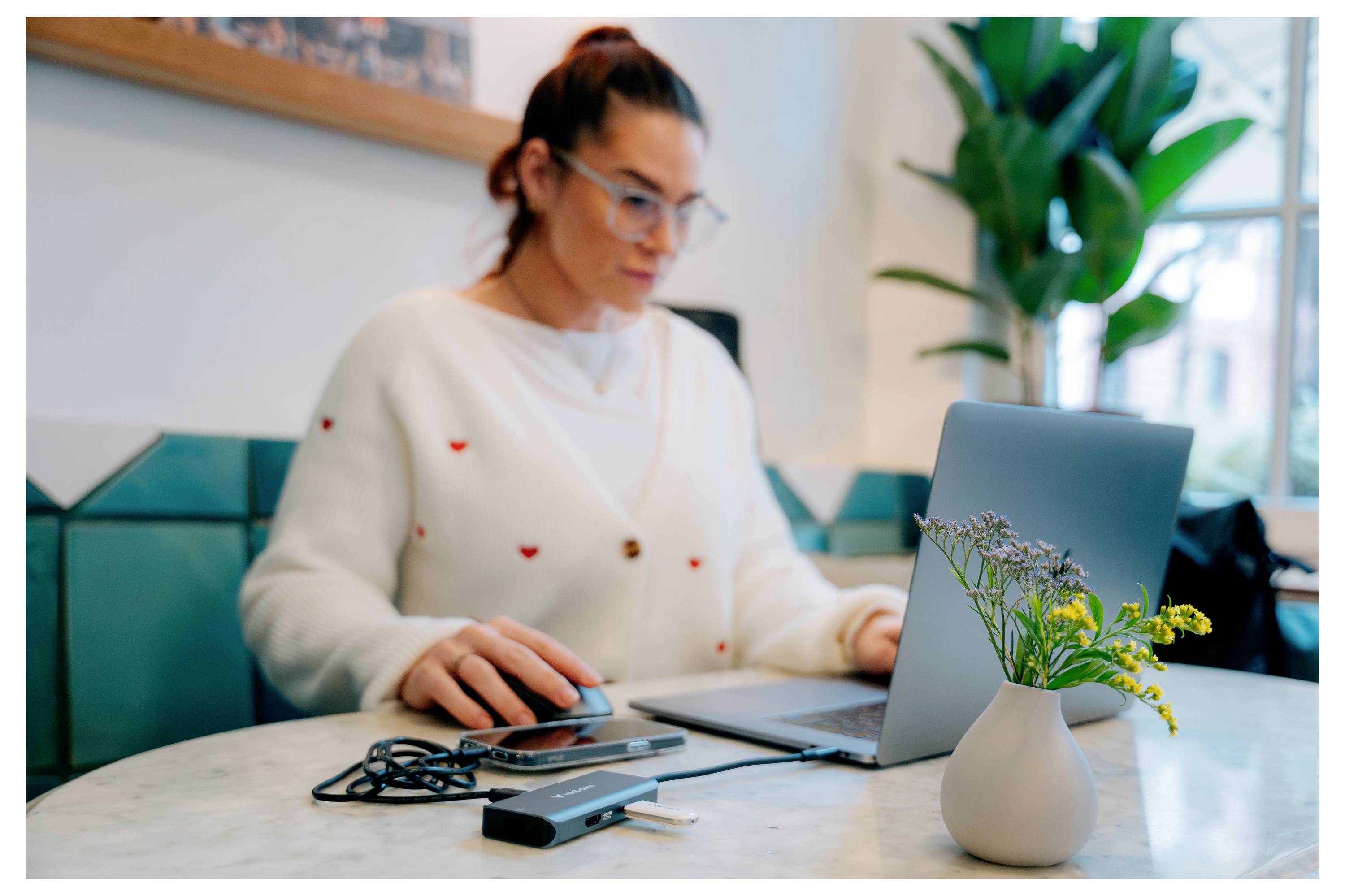 Eine Frau arbeitet an einem Laptop an einem Tisch in einem hellen Büro, mit einem Tablet, Kabeln und einer kleinen Vase mit Blumen in der Nähe.