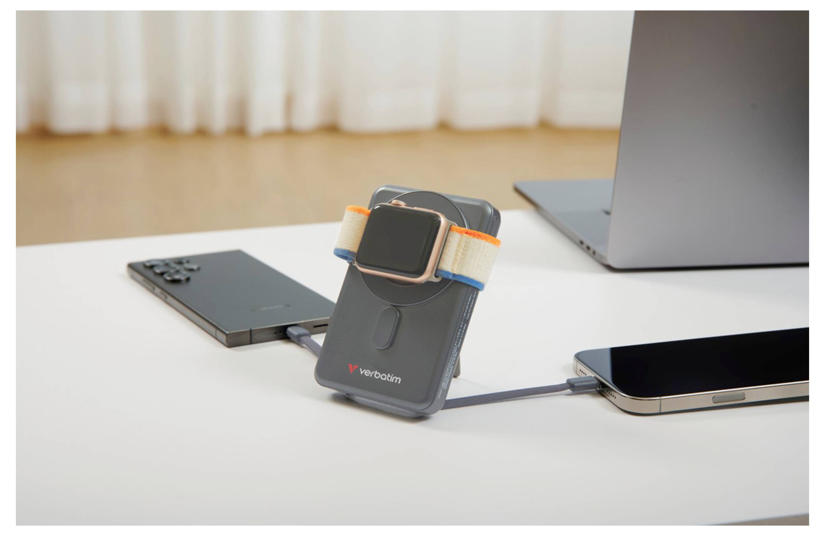 Eine Smartwatch wird auf einer tragbaren Powerbank aufgeladen, verbunden mit zwei Smartphones auf einem weißen Schreibtisch, mit einem Laptop im Hintergrund.