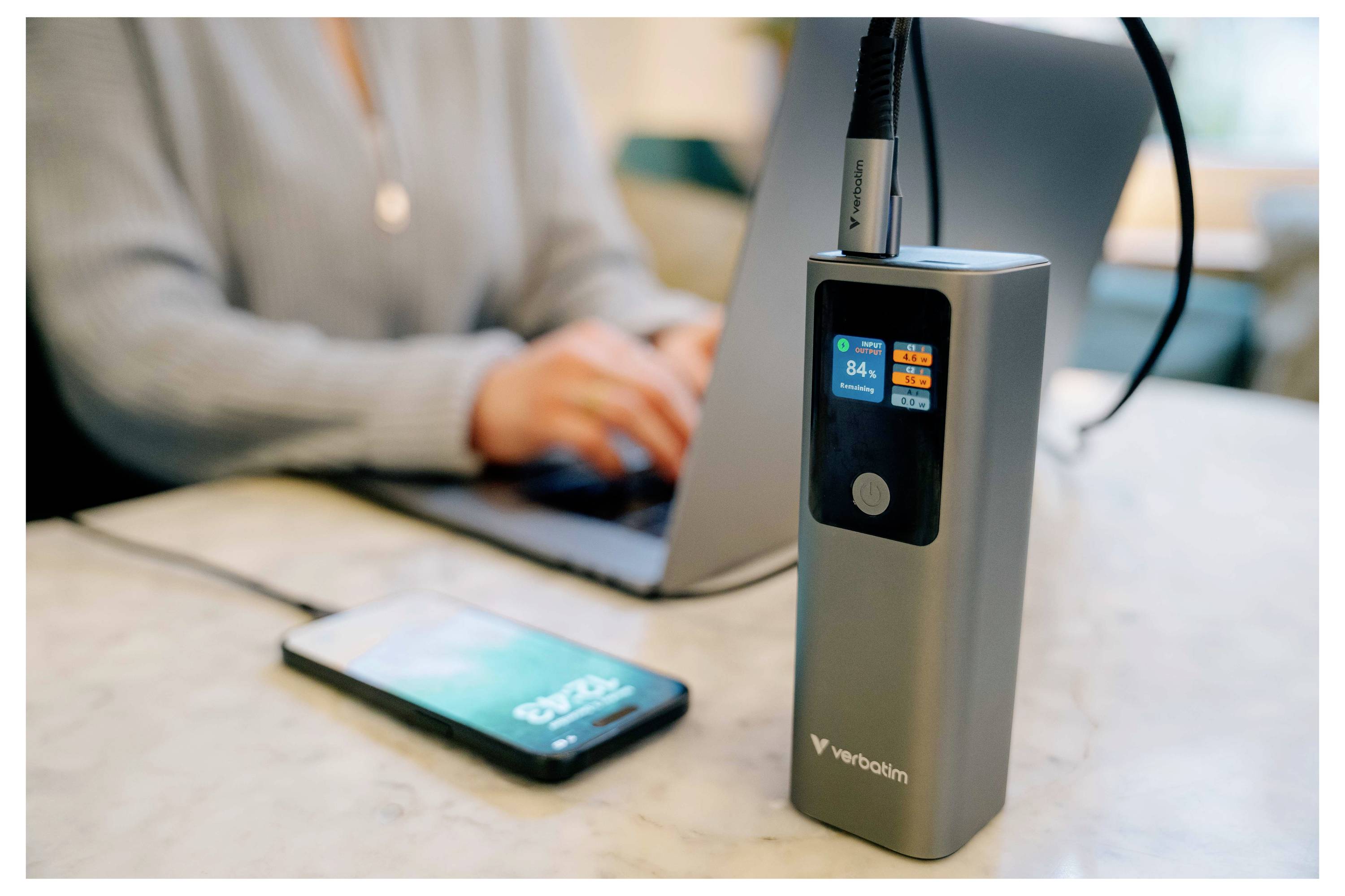 Tragbares Akkupack mit digitaler Ladeanzeige, das mit einem auf einem Schreibtisch aufladenden Telefon verbunden ist, während eine Person im Hintergrund einen Laptop benutzt.