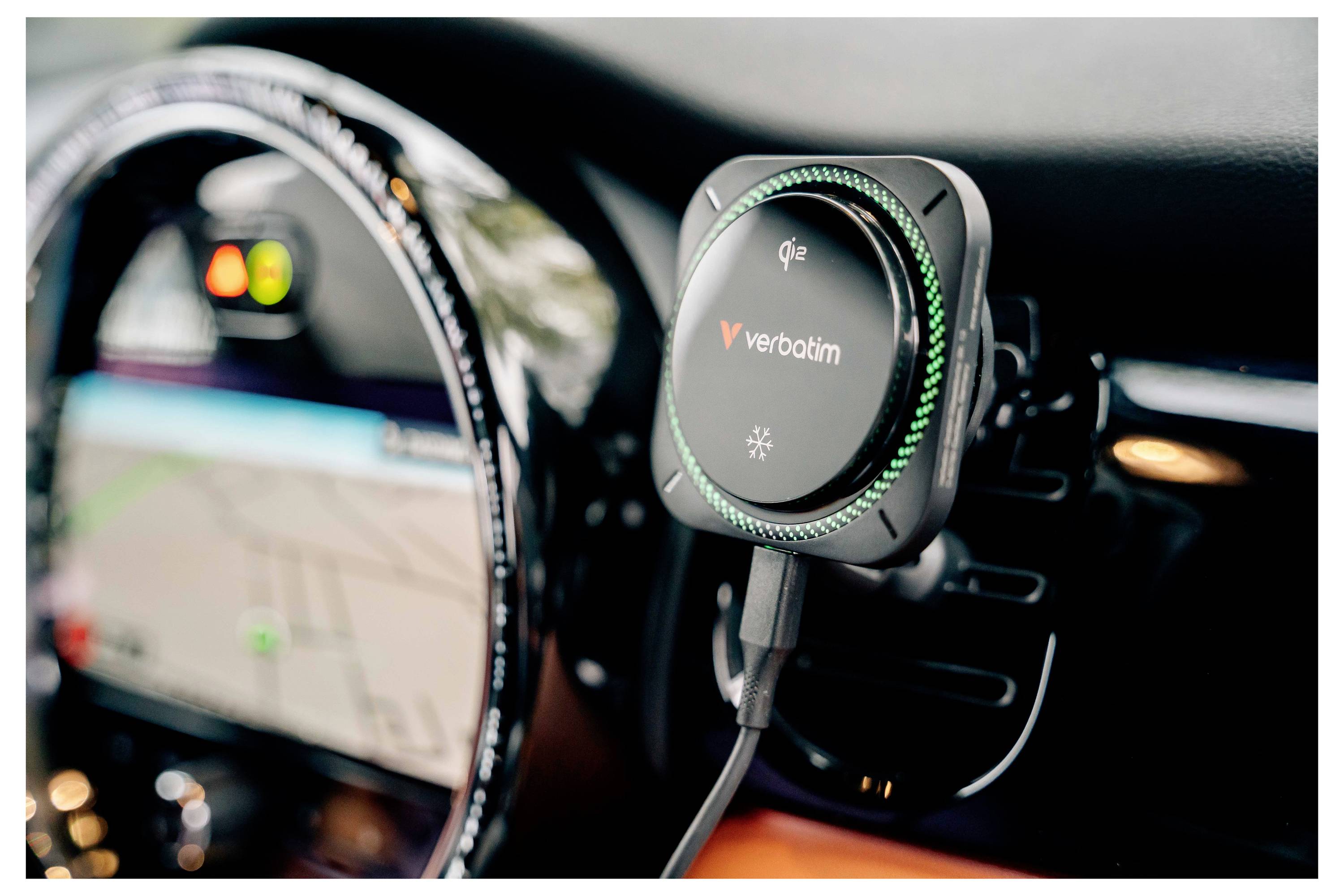 Ein Verbatim Qi-Ladegerät für kabellose Telefone, das auf einem Armaturenbrett eines Autos montiert ist, mit einem angeschlossenen Kabel und dem verschwommenen Kombiinstrument im Hintergrund.
