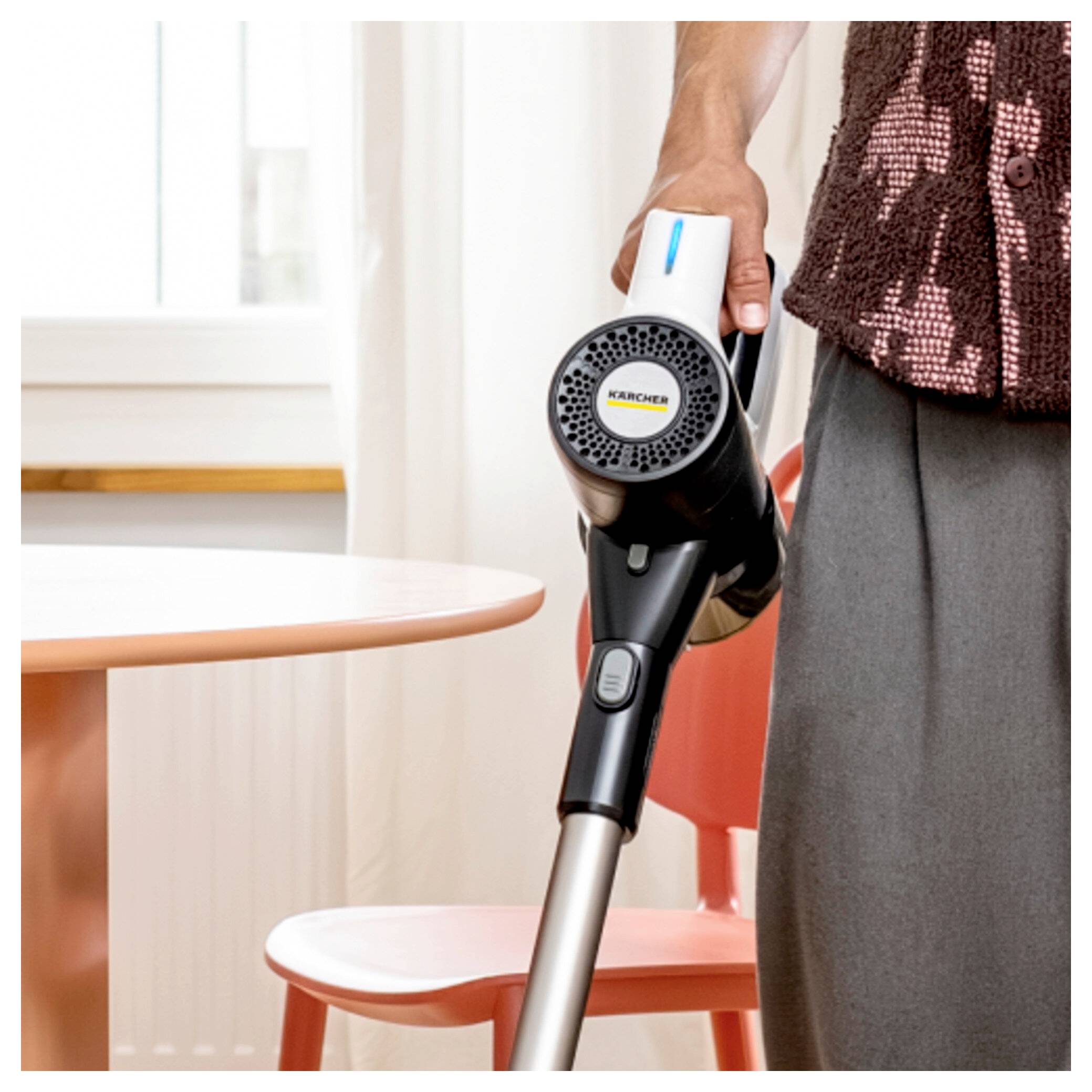 Eine Person hält einen tragbaren Staubsauger neben einem roten Tisch und Stuhl in einem hellen Raum. Das Gerät wird teilweise erfasst.