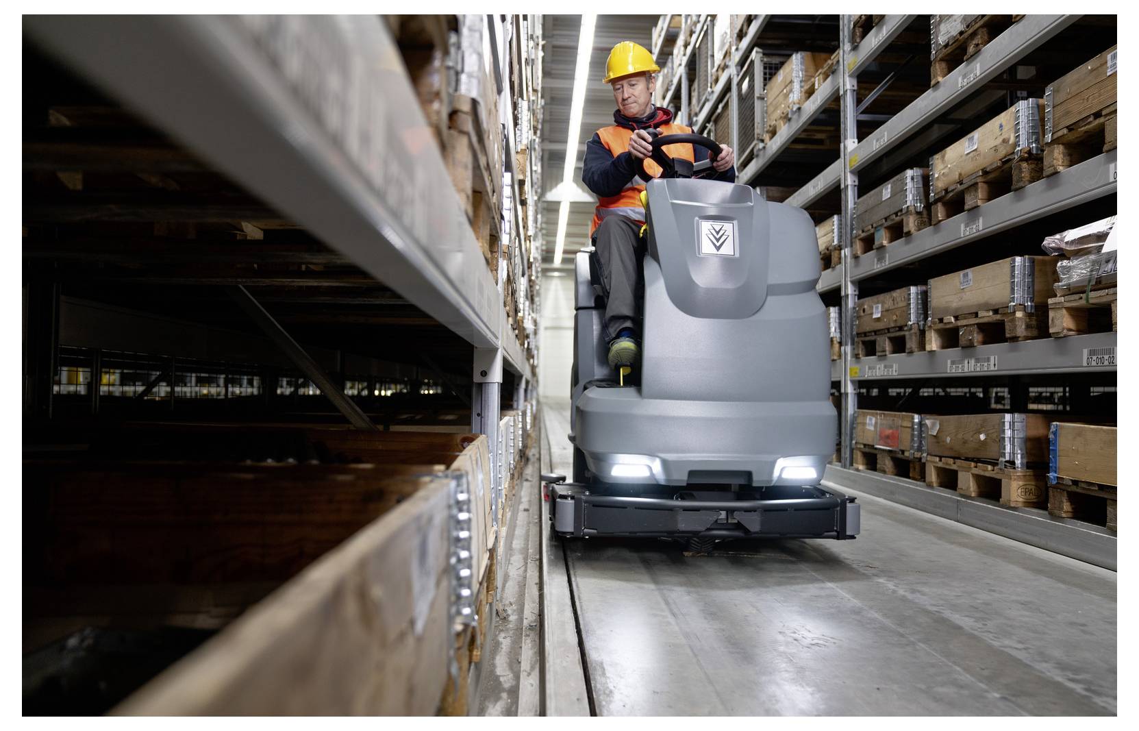 Ein Arbeiter mit Helm bedient eine industrielle Bodenschrubbermaschine in einem Lagergangsystem, das von Regalen voller Kartons umgeben ist.