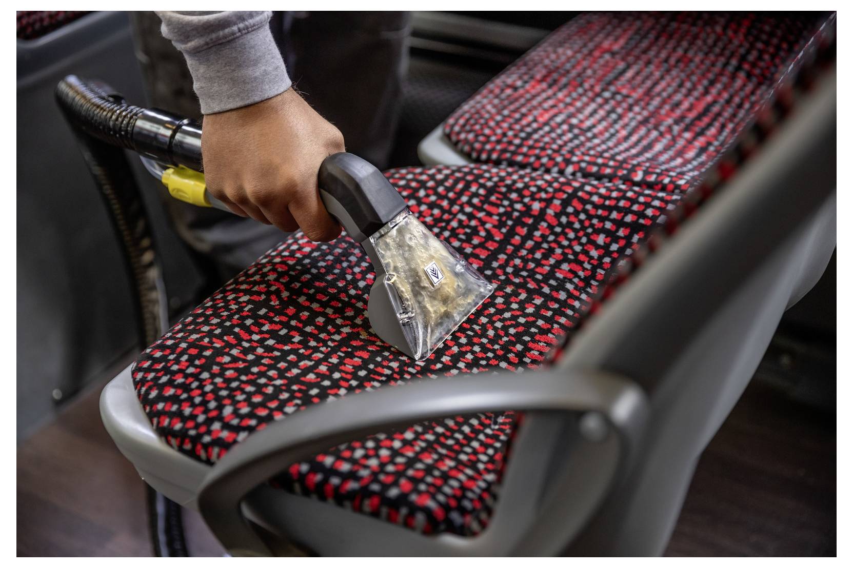 Eine Person benutzt einen Staubsauger, um den rot und schwarz gemusterten Stoffsitz eines Fahrzeugs zu reinigen.