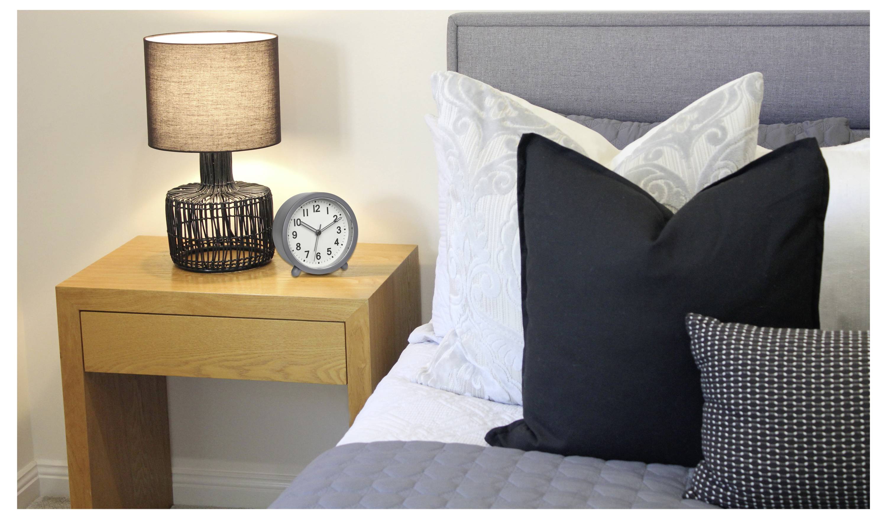 Schlafzimmerszene mit einem Nachttisch, auf dem eine Lampe und eine Uhr stehen, neben einem Bett mit einer grauen Steppdecke und weißen und schwarzen Kissen.