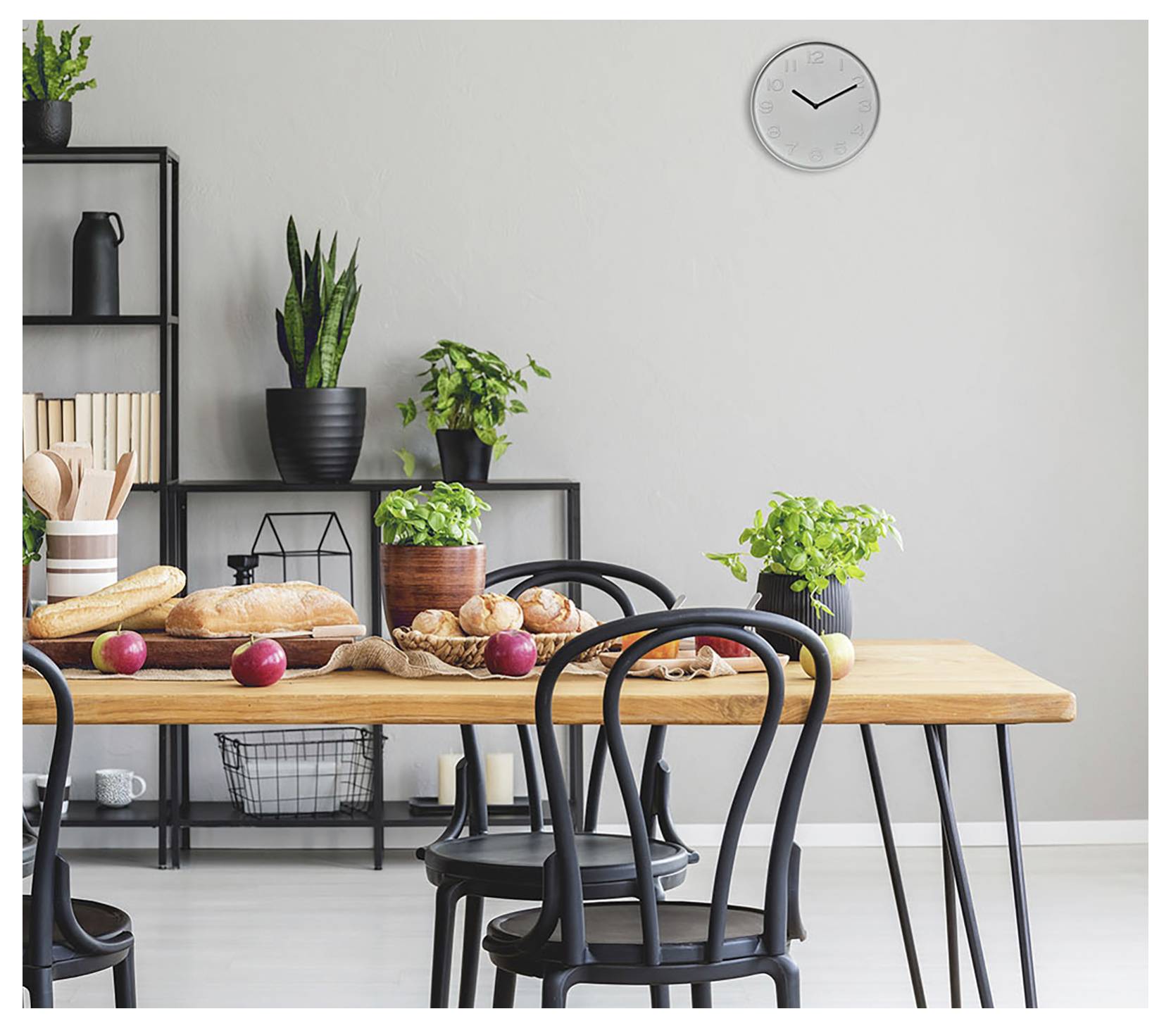 Ein modernes Esszimmer mit einem Holztisch, der mit Brot, Äpfeln und Pflanzen eingedeckt ist. Schwarze Stühle umgeben den Tisch. Eine Uhr und Regale sind sichtbar.
