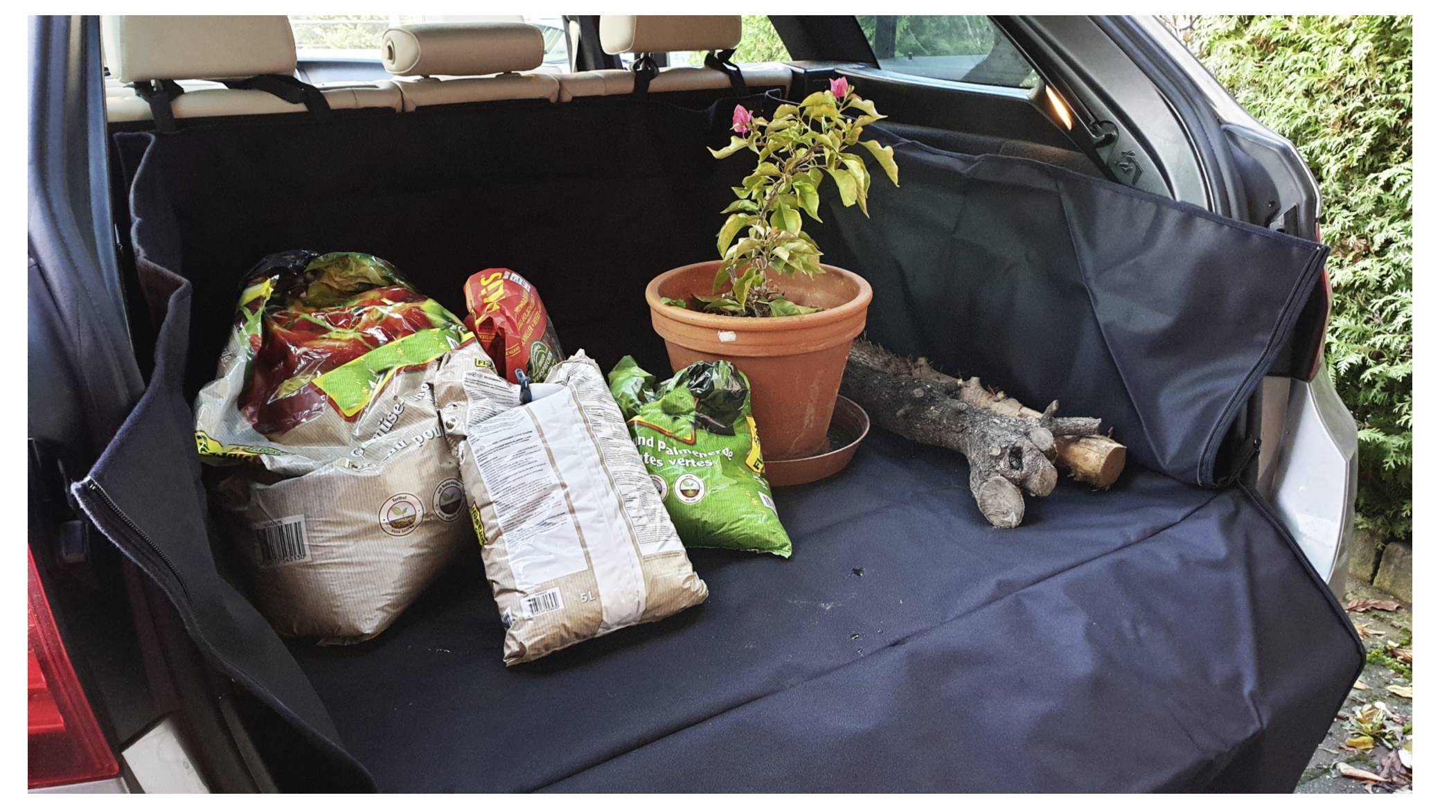 „Mehrere Säcke Erde, eine Topfpflanze mit rosa Blüten und Baumäste sind im offenen Kofferraum eines Autos angeordnet.