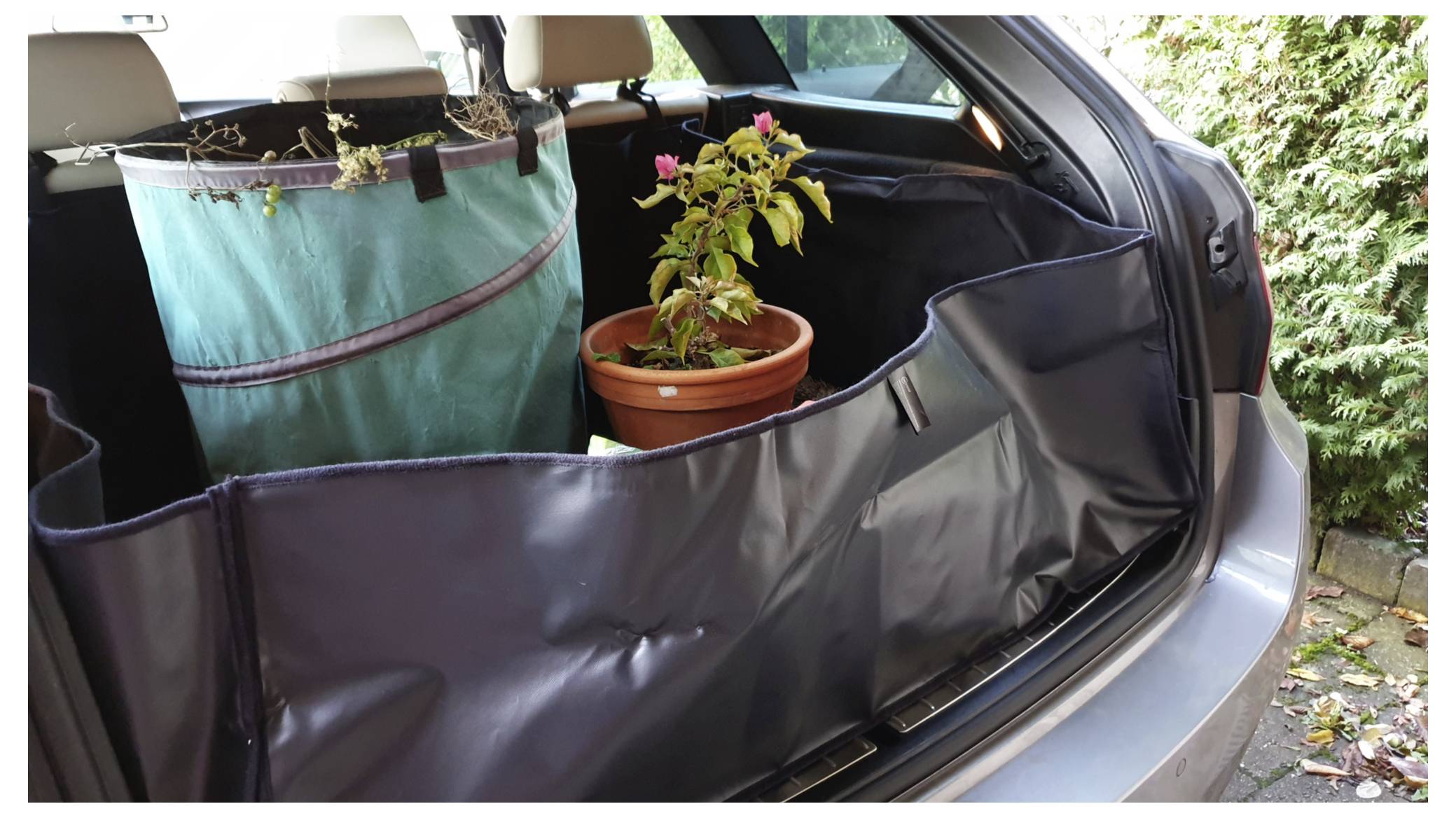 Kofferraum mit einer Topfpflanze und einem großen grünen Stoffbehälter. Der Kofferraum ist mit einer schützenden schwarzen Abdeckung ausgekleidet.