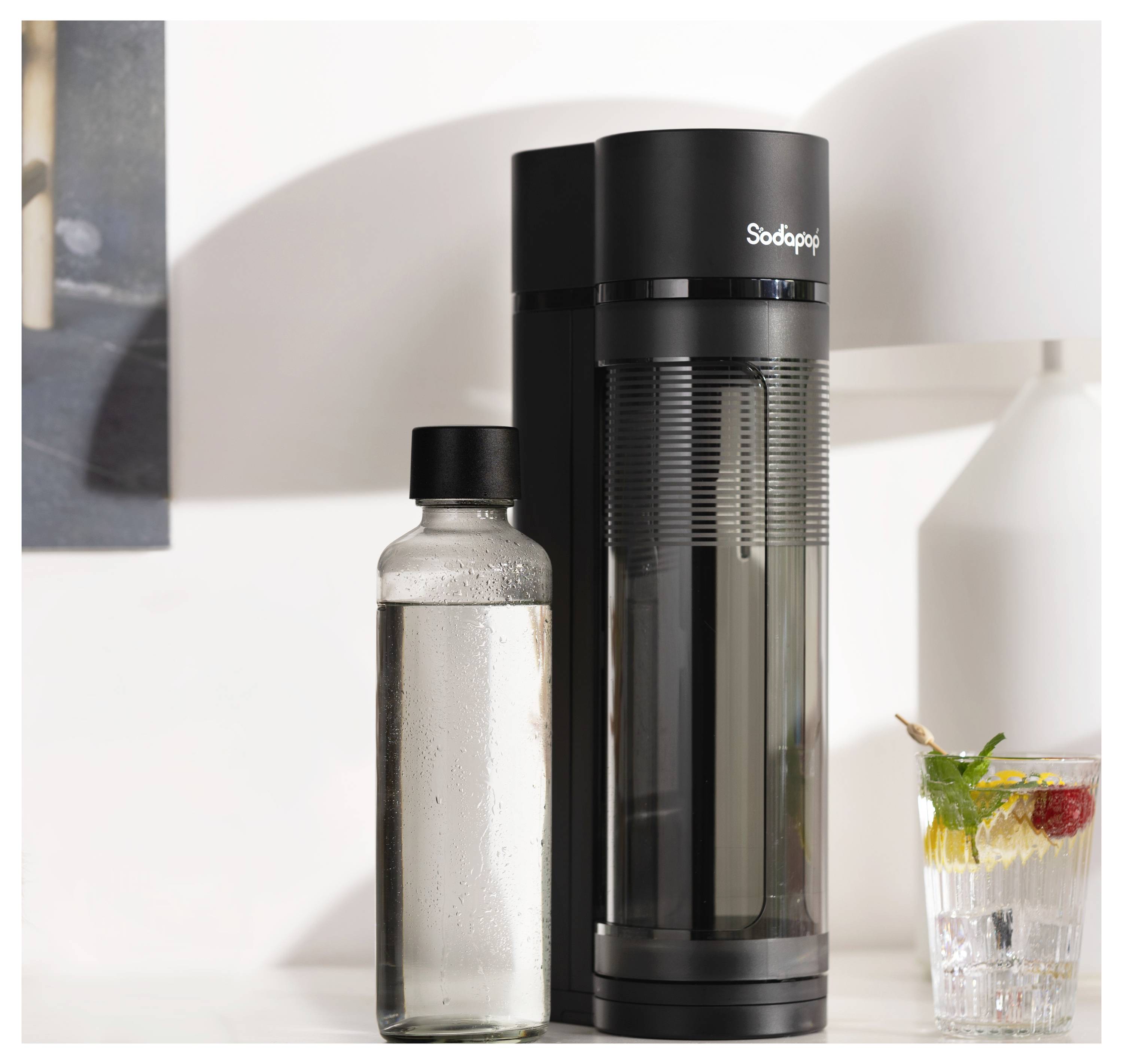 Ein schwarzer Wassersprudler steht auf einer Arbeitsplatte neben einer durchsichtigen Flasche gefüllt mit Wasser und einem Glas mit sprudelndem Getränk, garniert mit Minze und Obst.