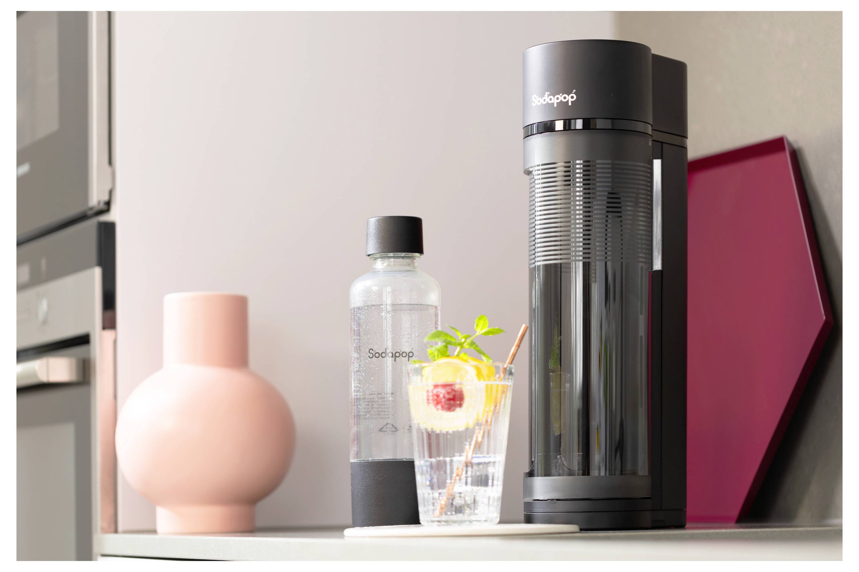 Ein schwarzer Wassersprudler auf einer Küchentheke neben einer Flasche und einem Glas mit gekühltem Fruchtwasser.