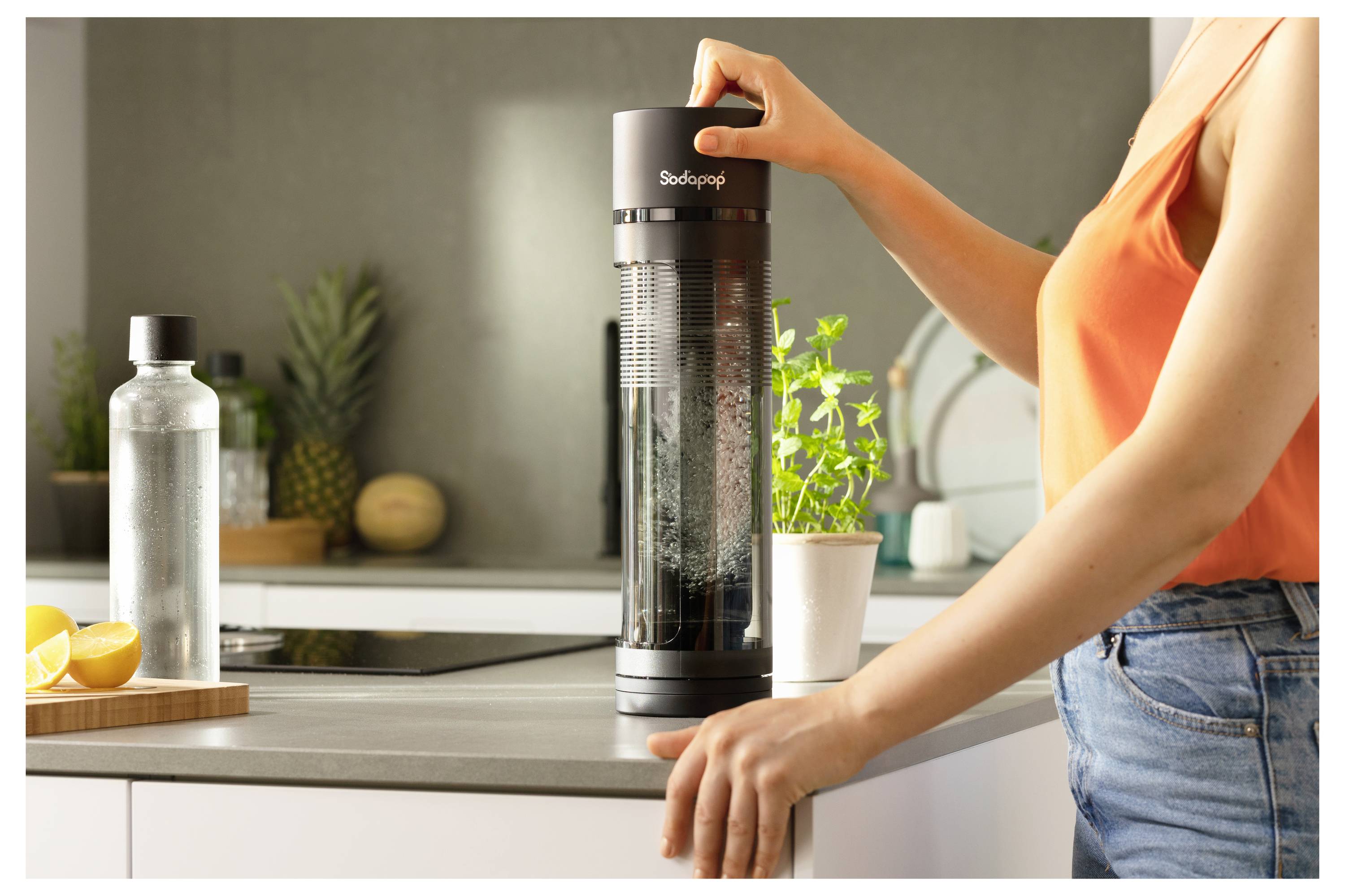 Eine Person in einem orangefarbenen Oberteil benutzt einen Sodabereiter auf einer Küchentheke. Eine Wasserflasche, Zitronen und frische Kräuter befinden sich in der Nähe.