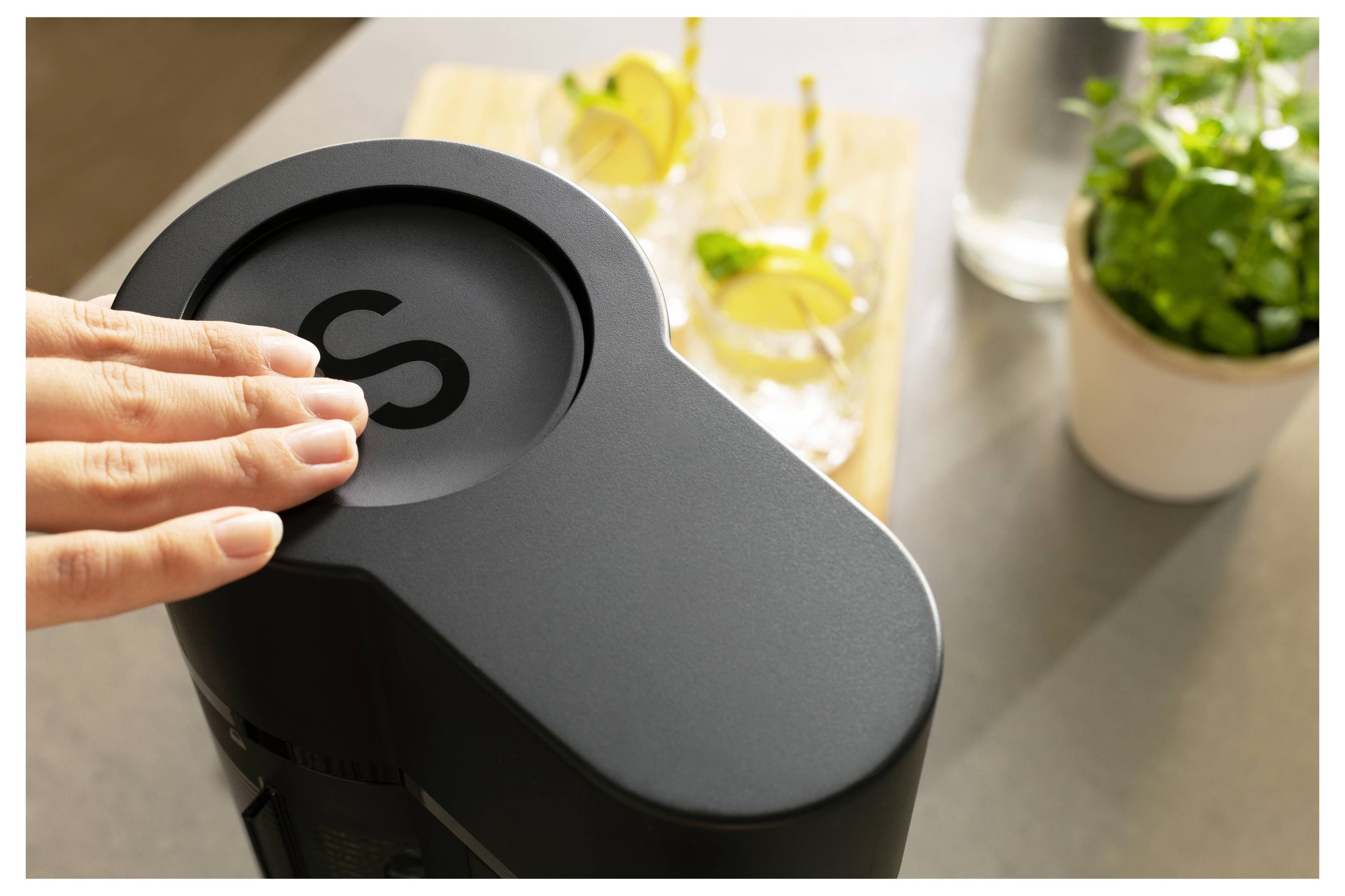 Eine Hand drückt die obere Taste eines modernen Küchengeräts, während zwei Getränke und eine Pflanze im Hintergrund zu sehen sind, was auf die Zubereitung von Getränken hindeutet.