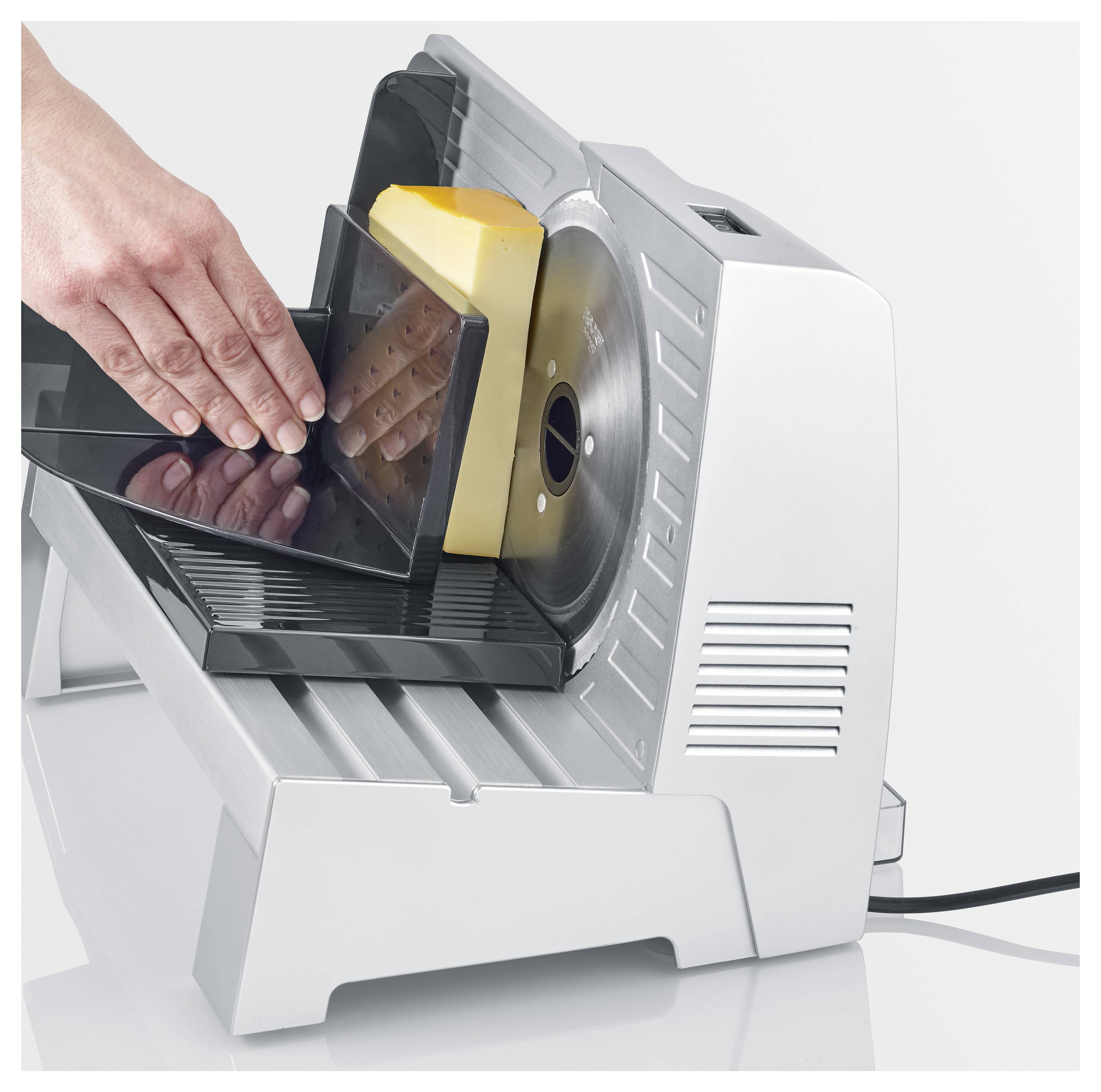 Eine Person nutzt einen elektrischen Käsehobel, um einen Käseblock zu schneiden. Der silberfarbene Körper des Hobels und die Sicherheitsblende sind deutlich sichtbar.