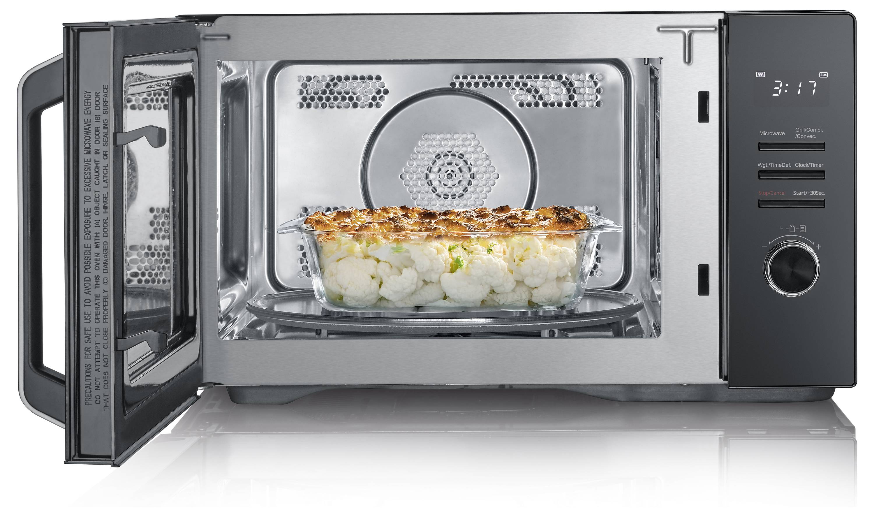 Eine offene Mikrowelle mit einer Glasschüssel im Inneren, die weiße und gebräunte Lebensmittel enthält, wahrscheinlich einen Blumenkohl-Auflauf, vor einem silbernen Innenraum.