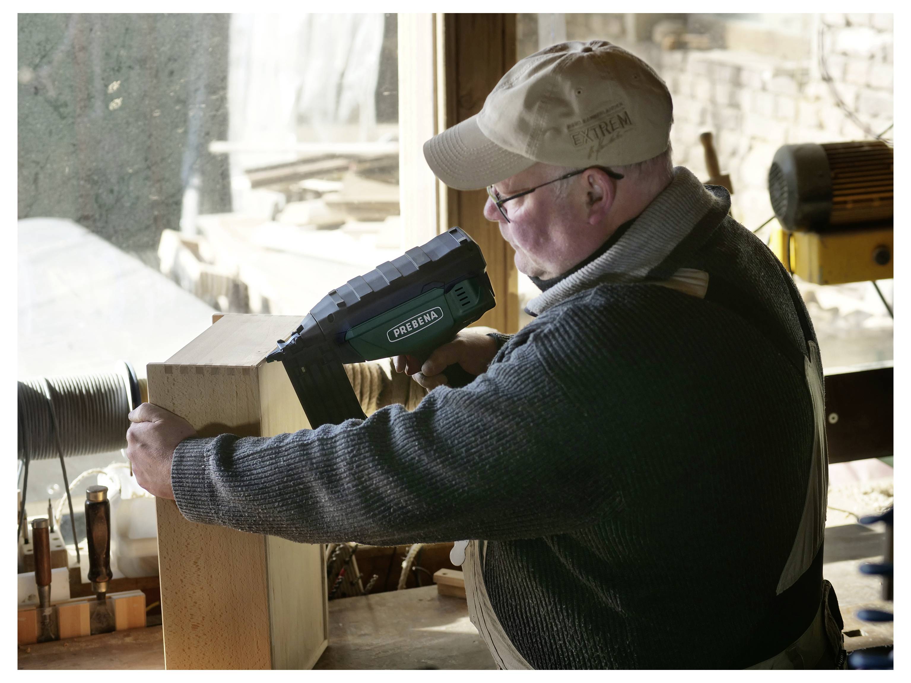 Ein Mann, der eine Kappe und eine Brille trägt, benutzt eine Nagelschussmaschine, um Holzteile in einer Werkstatt mit Werkzeugen und Holz im Hintergrund zusammenzusetzen.