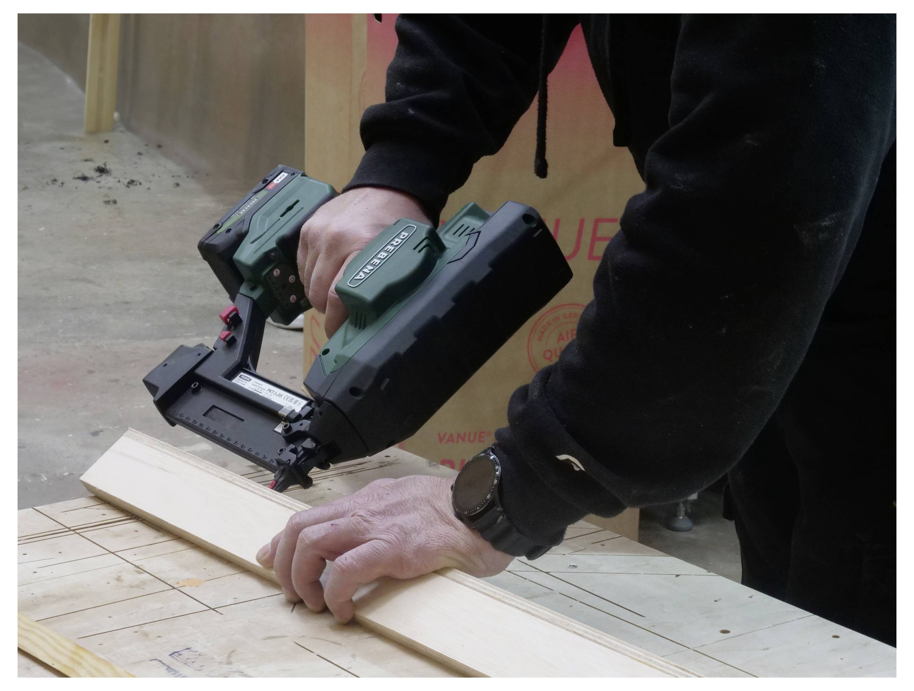 Eine Person benutzt eine grüne Nagelschussmaschine, um ein Stück Holz auf einem Tisch zu befestigen, trägt einen schwarzen Hoodie und eine Uhr in einer Werkstattsituation.