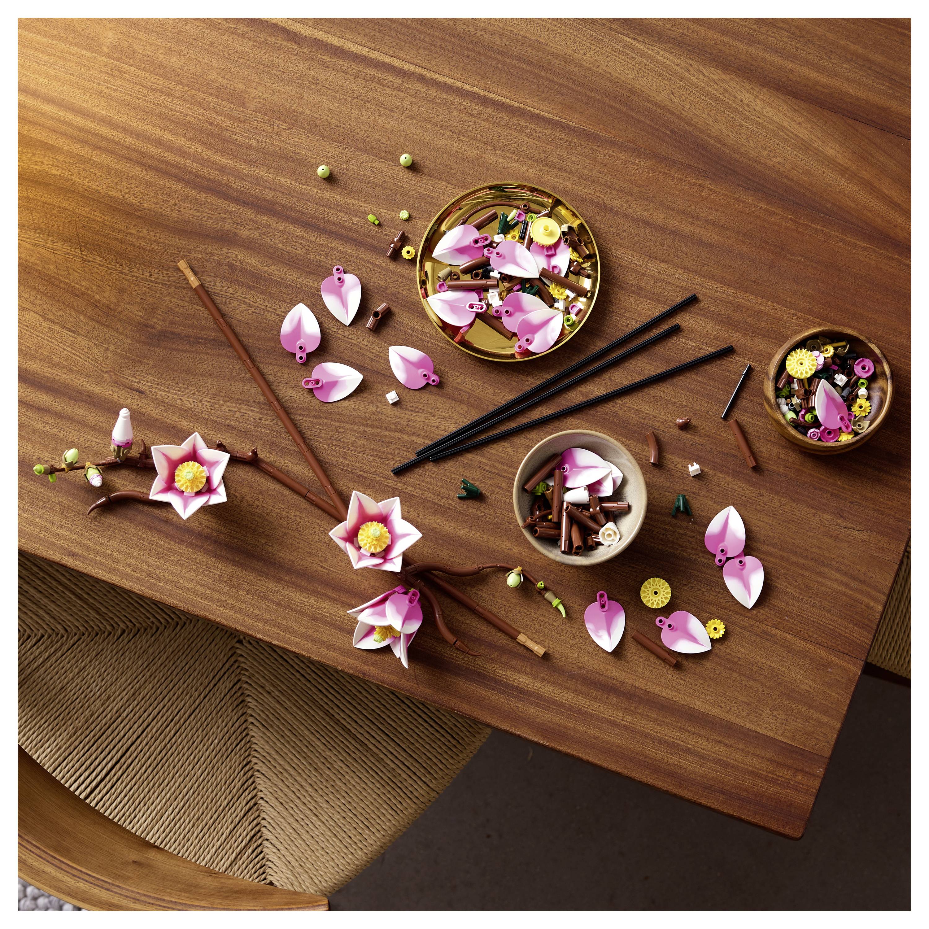 Holztisch mit zusammengesetzten und zerlegten rosa- und weißfarbenen LEGO-Blumenteilen, Essstäbchen und Schüsseln mit Blütenteilen.