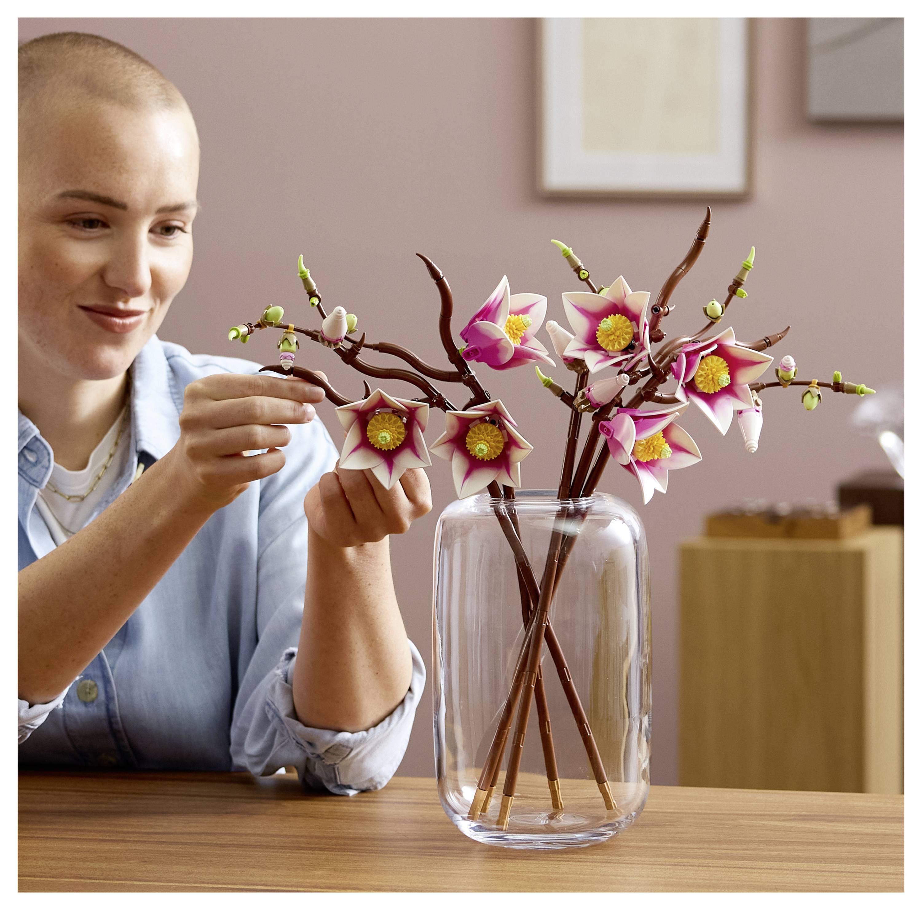 Eine Person arrangiert zarte rosa und gelbe Blüten mit Knospen auf Zweigen in einer klaren Glasvase und schafft damit eine schlichte, aber elegante Dekoration.