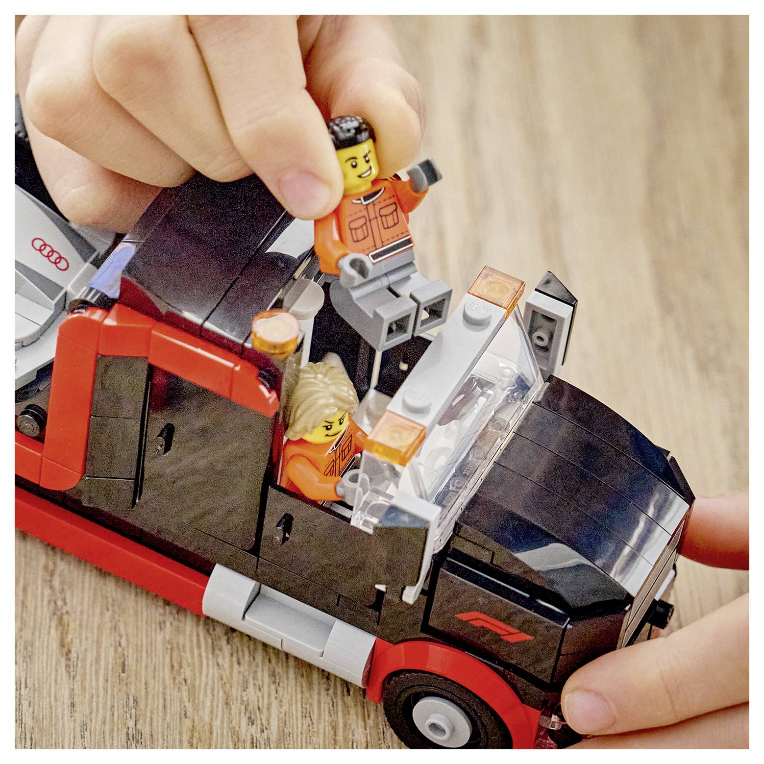 Zwei Hände spielen mit kleinen Figuren in einem Spielzeuglastauto-Modell auf einer Holzoberfläche.