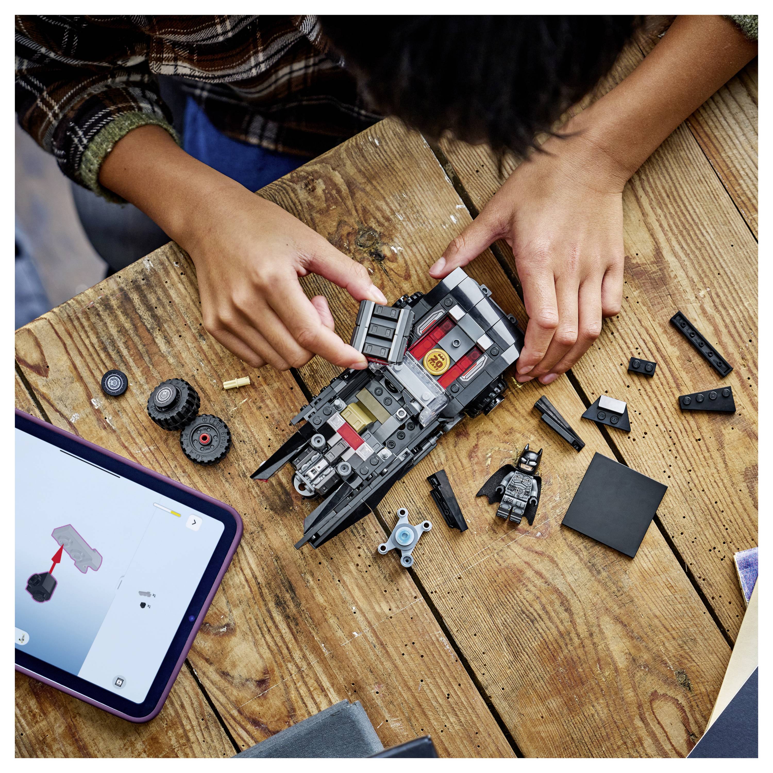 Eine Person, die ein LEGO-Batmobil auf einem Holztisch zusammenbaut, mit Anleitung auf einem Tablet und verschiedenen LEGO-Steinen rundherum verstreut.
