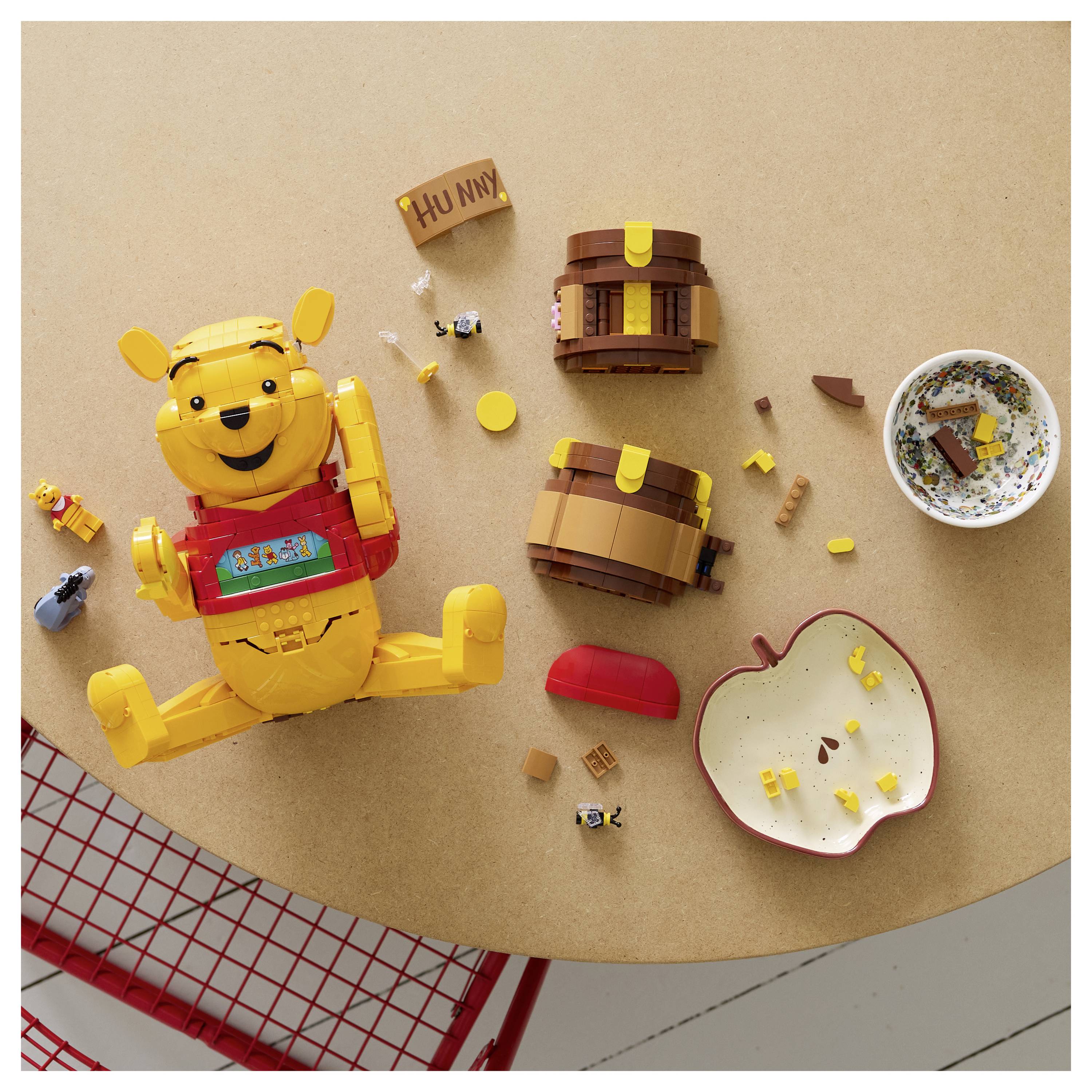 Ein LEGO-Set mit einem Bären und einem Honigopf, umgeben von verstreuten Teilen auf einem Tisch, neben einer leeren Schüssel und einem roten apfelförmigen Behälter.
