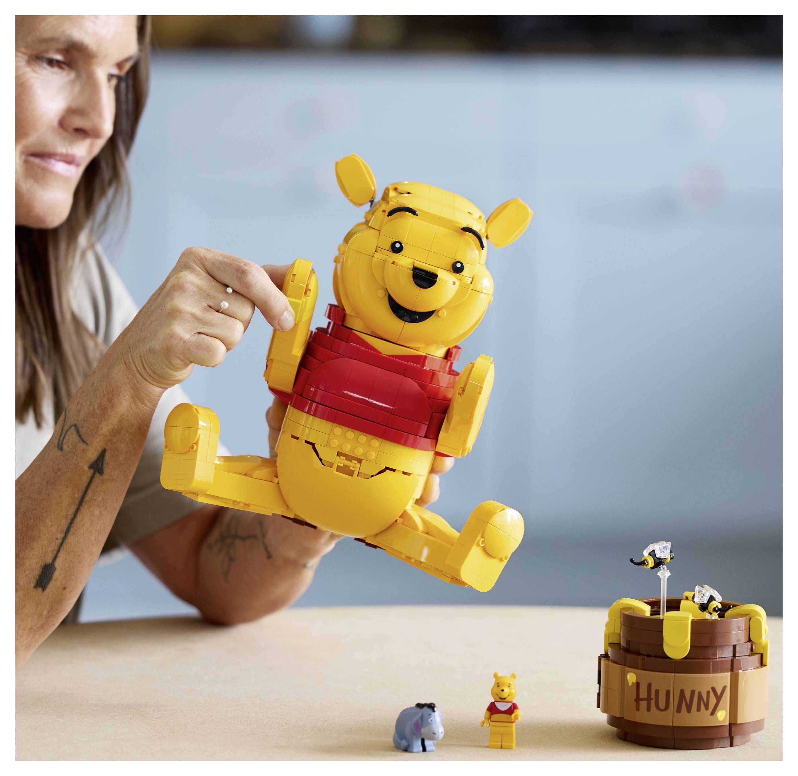 Eine Person interagiert mit einer großen, zusammengebauten Spielfigur, die einem Bären in Rot ähnelt. In der Nähe befinden sich kleinere Bären- und Eselspielzeuge sowie ein „Hunny