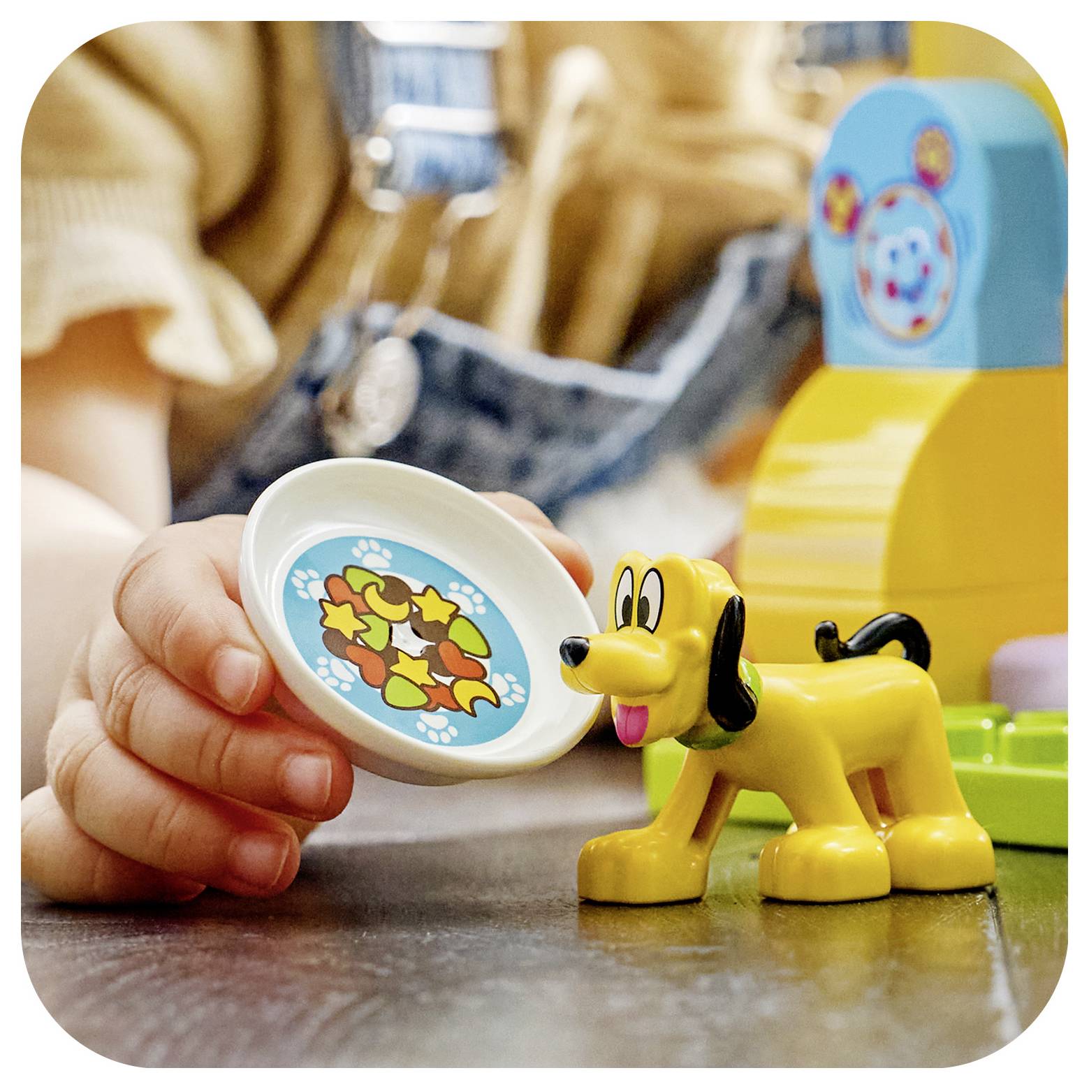 Die Hand eines Kindes hält einen Spielzeugteller mit einem Hundeknochen-Muster neben einem kleinen gelben Kunststoffhund auf einem Tisch, mit Spielzeugen im Hintergrund.