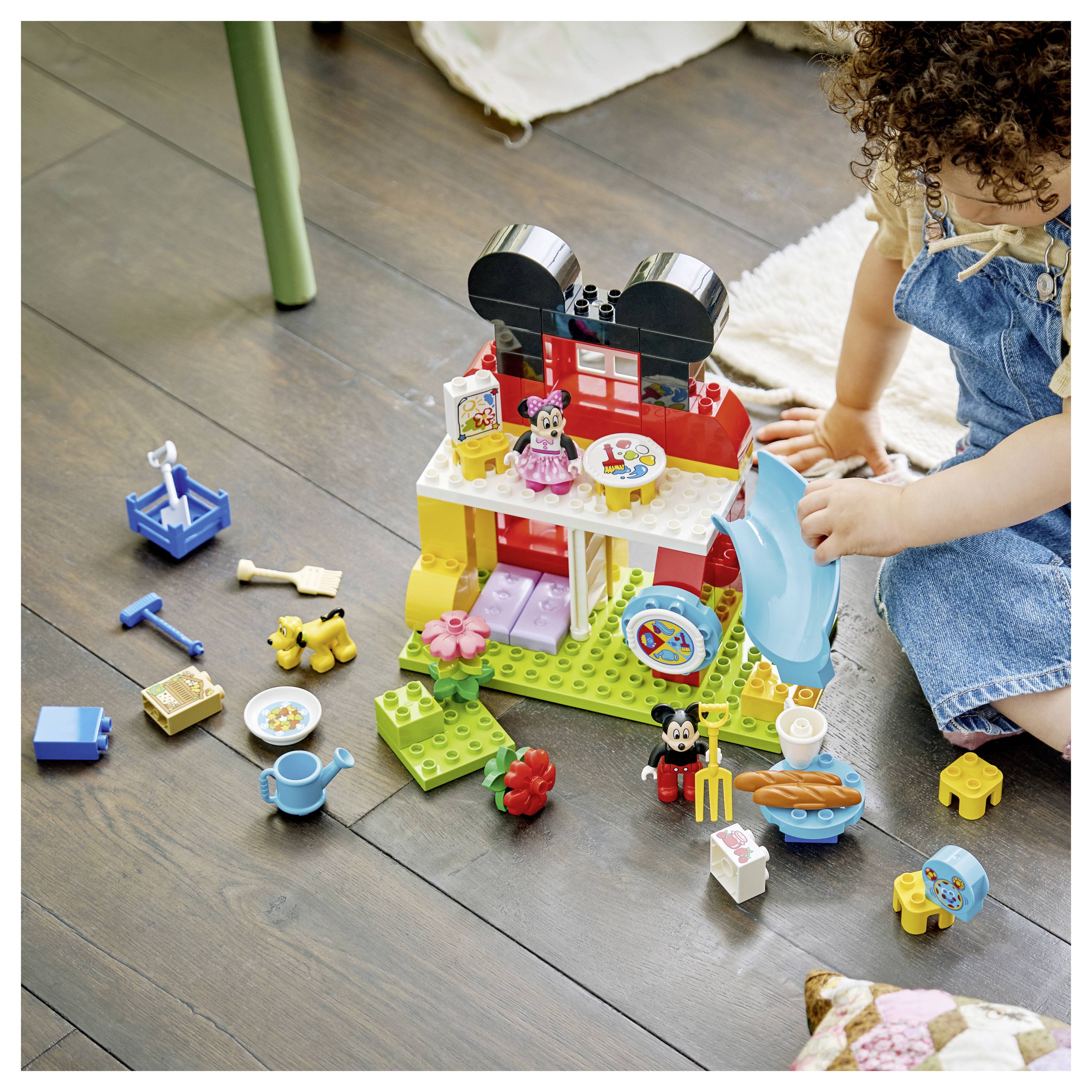 Ein Kind spielt mit Bausteinen und setzt ein farbenfrohe Hausset mit verschiedenen kleinen Zubehörteilen auf einem Holzboden zusammen.