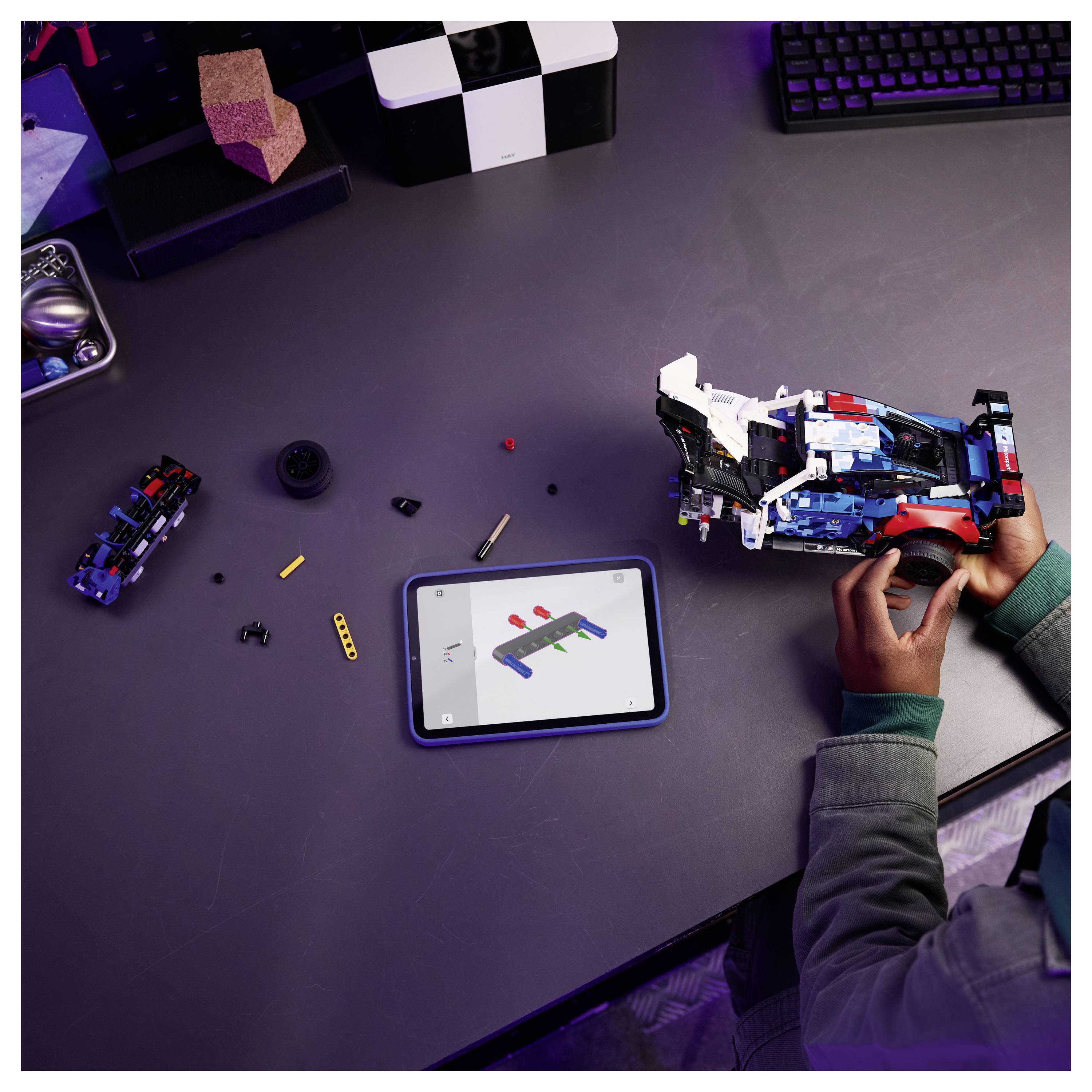 Eine Person, die ein LEGO-Modell mit einem Tablet zusammensetzt, auf dem Anweisungen angezeigt werden. Verschiedene LEGO-Teile und Werkzeuge sind auf dem Tisch verteilt.