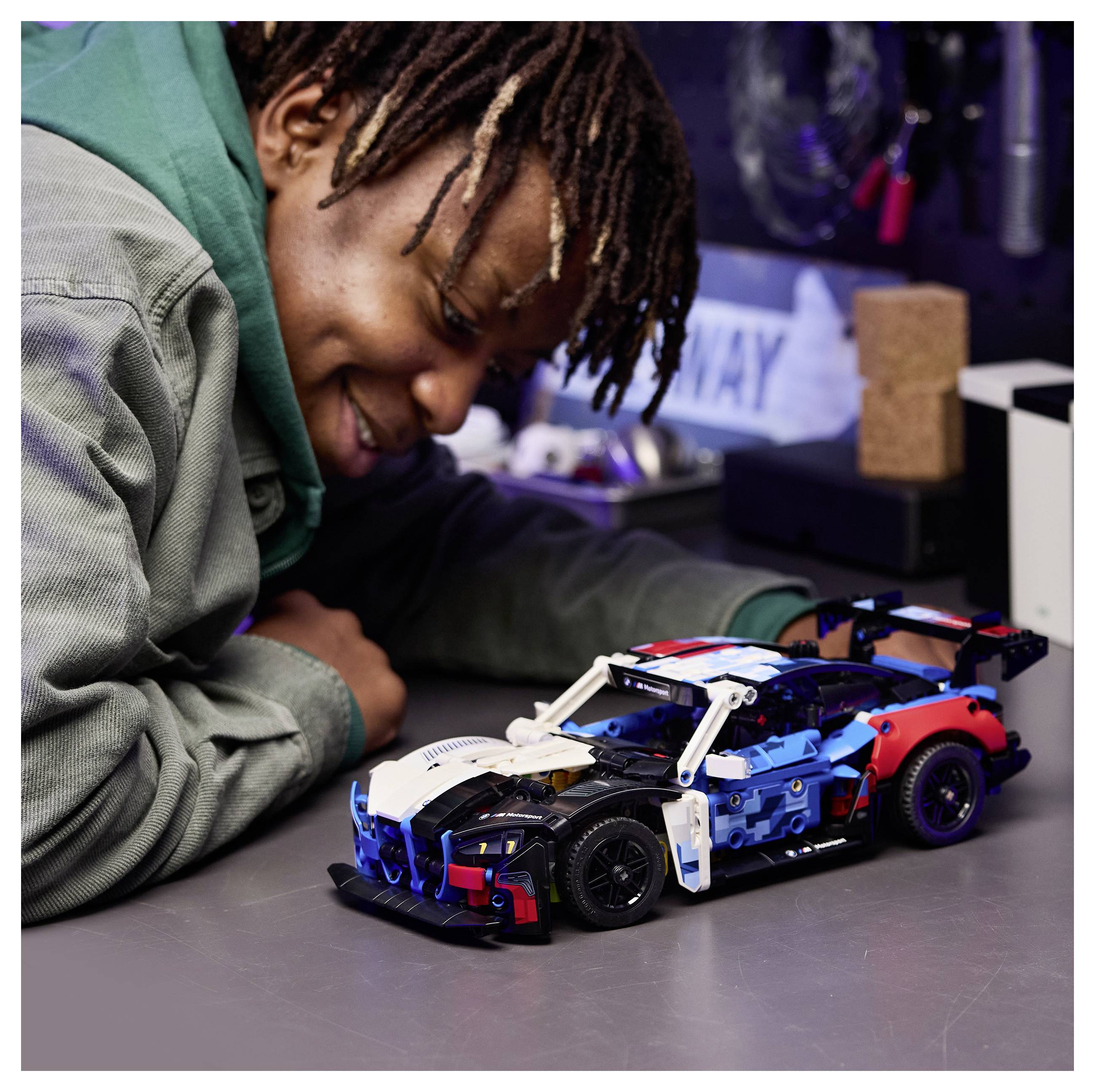 Eine Person, die lächelt und sich über ein detailliertes Spielzeug-Rennwagen-Modell auf einem Tisch beugt, was Freude und Konzentration in einer Werkstatt-Umgebung anzeigt.