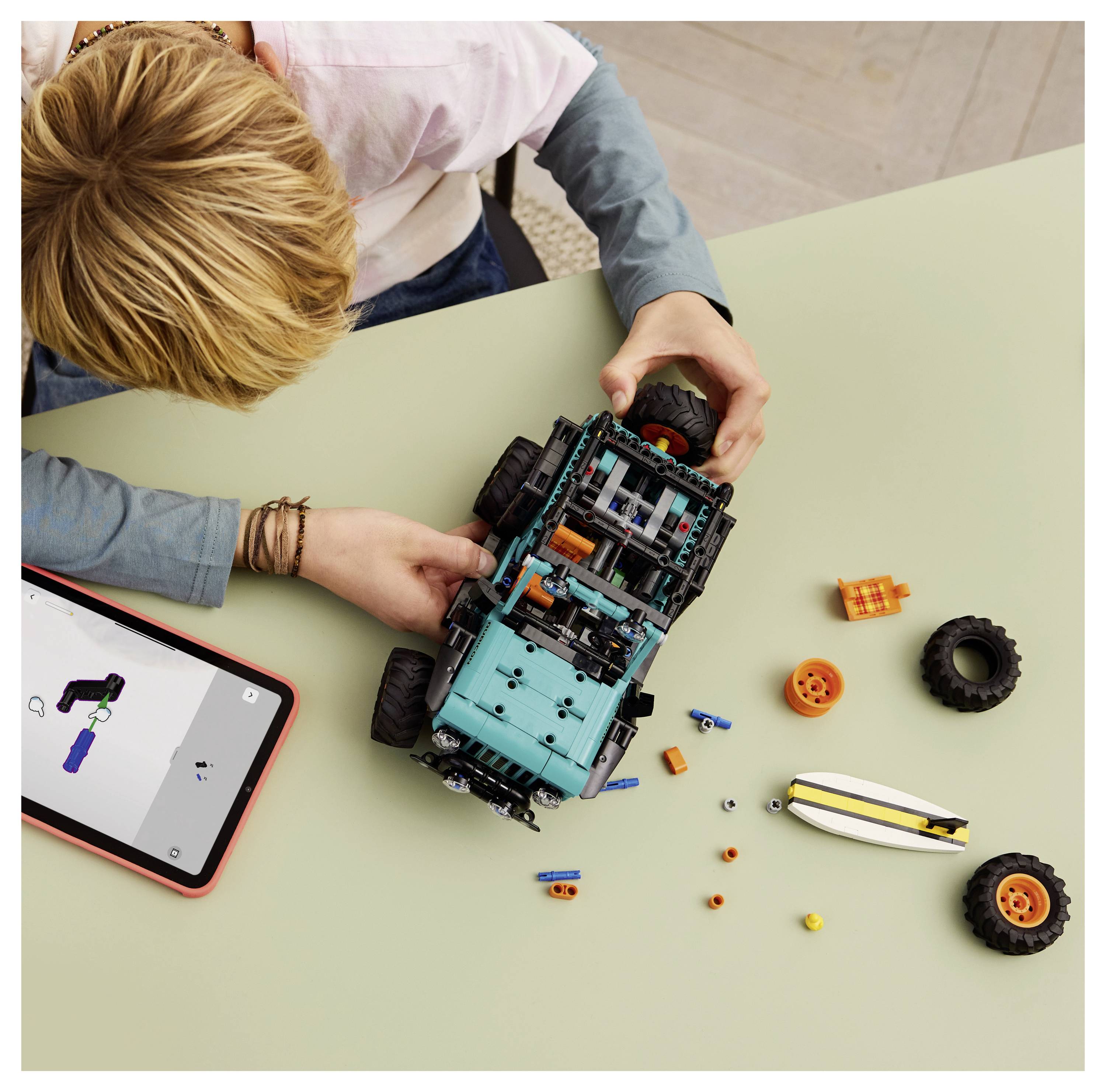 Ein Kind montiert ein blaues Spielzeugfahrzeug mit schwarzen Rädern und orange-blauen Komponenten auf einem Tisch. Ein Tablet zeigt Montageanweisungen an.