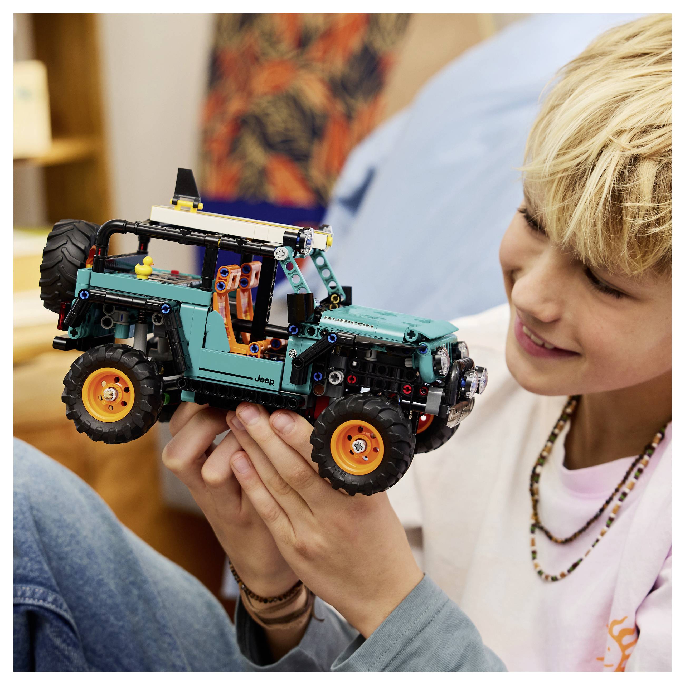 Eine Person lächelt, während sie einen detaillierten Spielzeug-Jeep mit leuchtend orangefarbenen Rädern hält und sein realistisches Design sowie seine komplexe Montage zur Schau stellt.