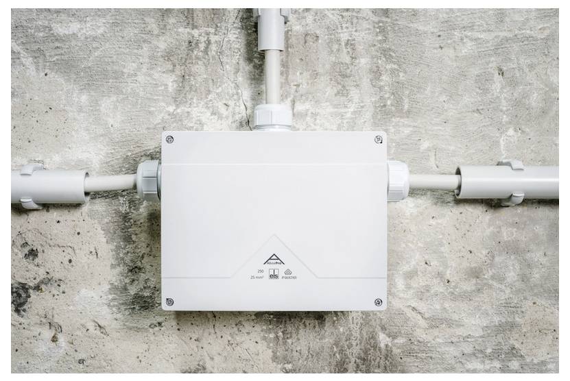 Weiße elektrische Verteilerdose, die an einer Betonwand montiert ist, mit Rohren, die von oben und von beiden Seiten eindringen.
