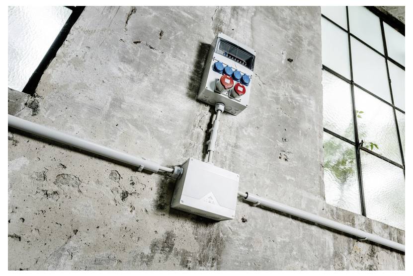 Industrielle Wand mit Elektroschutzschalterkasten, Steckdosenfeld und Rohrleitungen neben einem großen Fenster.