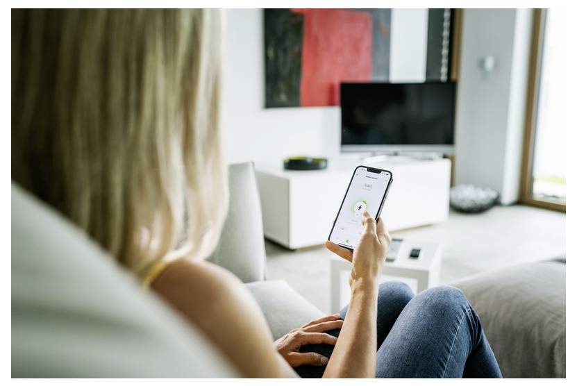 Frau auf einem Sofa, die ein Smartphone nutzt, um ein Smart-Home-Gerät in einem modernen Wohnzimmer mit einem Fernseher und einem Lautsprecher im Hintergrund zu steuern
