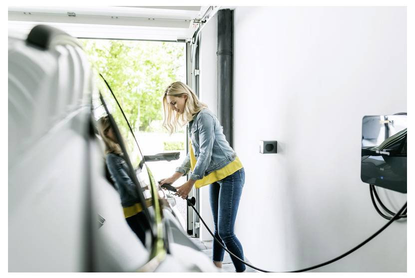Frau, die ein Elektroauto in einer Garage auflädt, das Kabel in das Fahrzeug einsteckt.